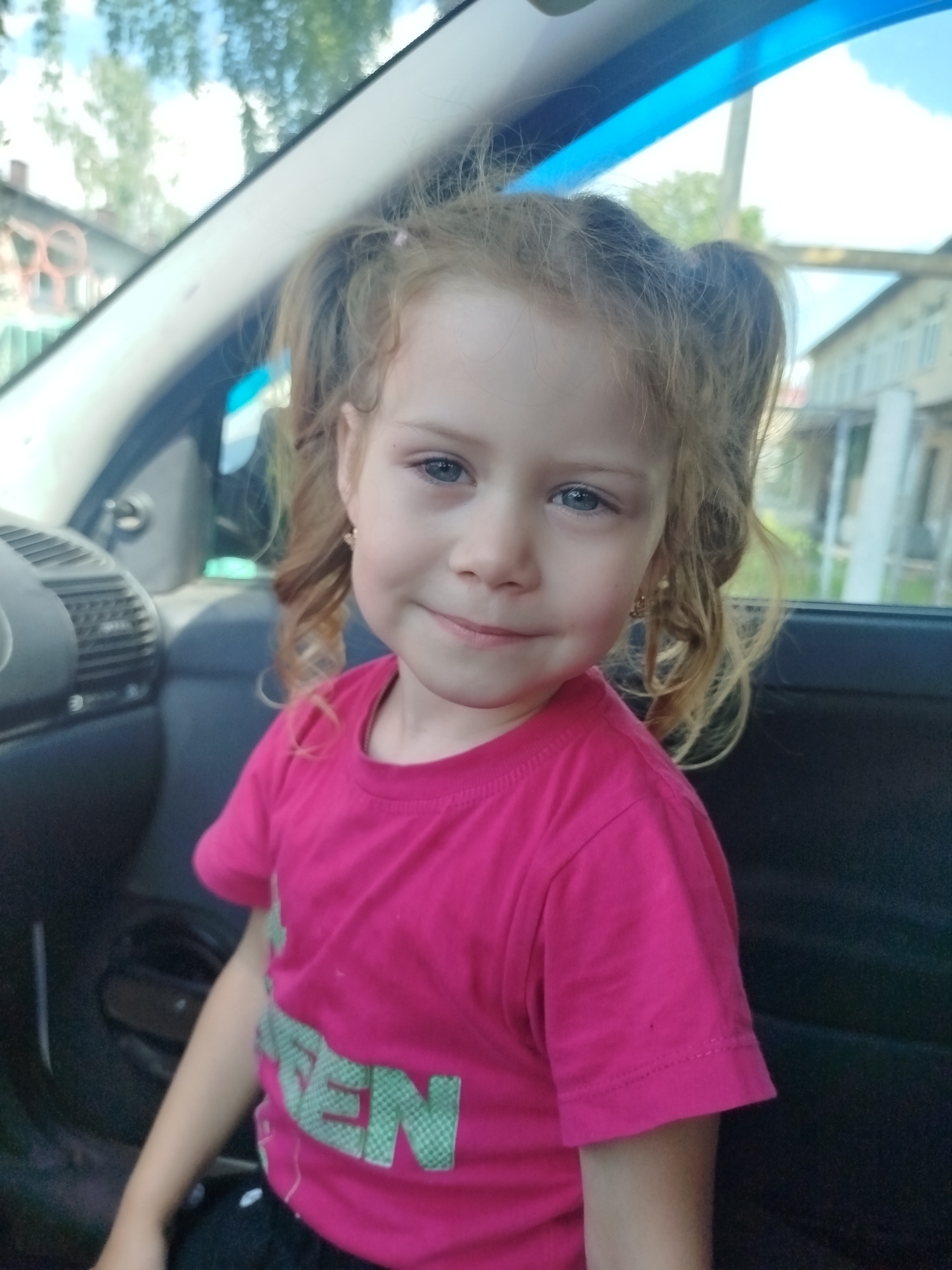 The image shows a young Caucasian girl with reddish-blonde hair in two pigtails, wearing a bright pink t-shirt with green lettering on it, sitting in the passenger seat of a car. The girl has blue eyes and is smiling slightly at the camera. Her hair is styled in curls. The interior of the car is visible, including a black dashboard with air vents and a dark-colored seat. Outside the car window, houses and trees can be seen under a bright sky. The scene appears to be in a rural or suburban setting. The car is likely parked in front of a residential building in Krasny Gulyai, Russia, based on the context provided.