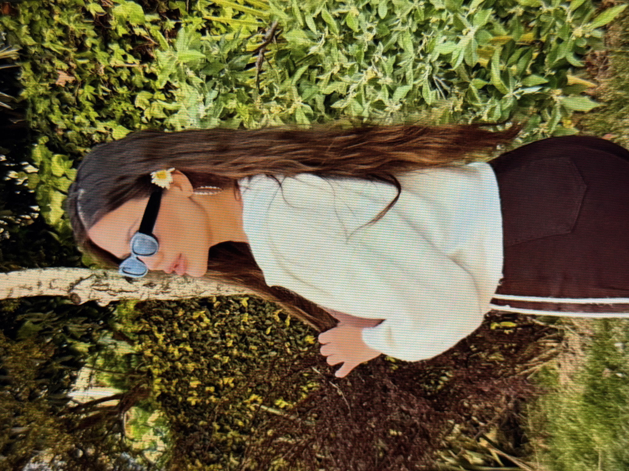 This media file captures a close-up, horizontally oriented, outdoor shot of a young person with long, dark brown hair, adorned with a small white flower tucked behind their ear. They are wearing dark sunglasses and a white long-sleeved top. The background consists of lush green foliage and grass, suggesting a natural, possibly garden or park setting. The person is positioned in a way that their body is partially obscured by the clothing they are wearing. Their head is turned to the side, with their face tilted downwards. A hint of a dark-colored garment, possibly pants or a skirt, is visible on the right side of the frame. The overall impression is one of a casual, relaxed moment outdoors, with the vibrant green of nature contrasting with the clothing. There is no visible text in the image. The lighting suggests it is daytime.