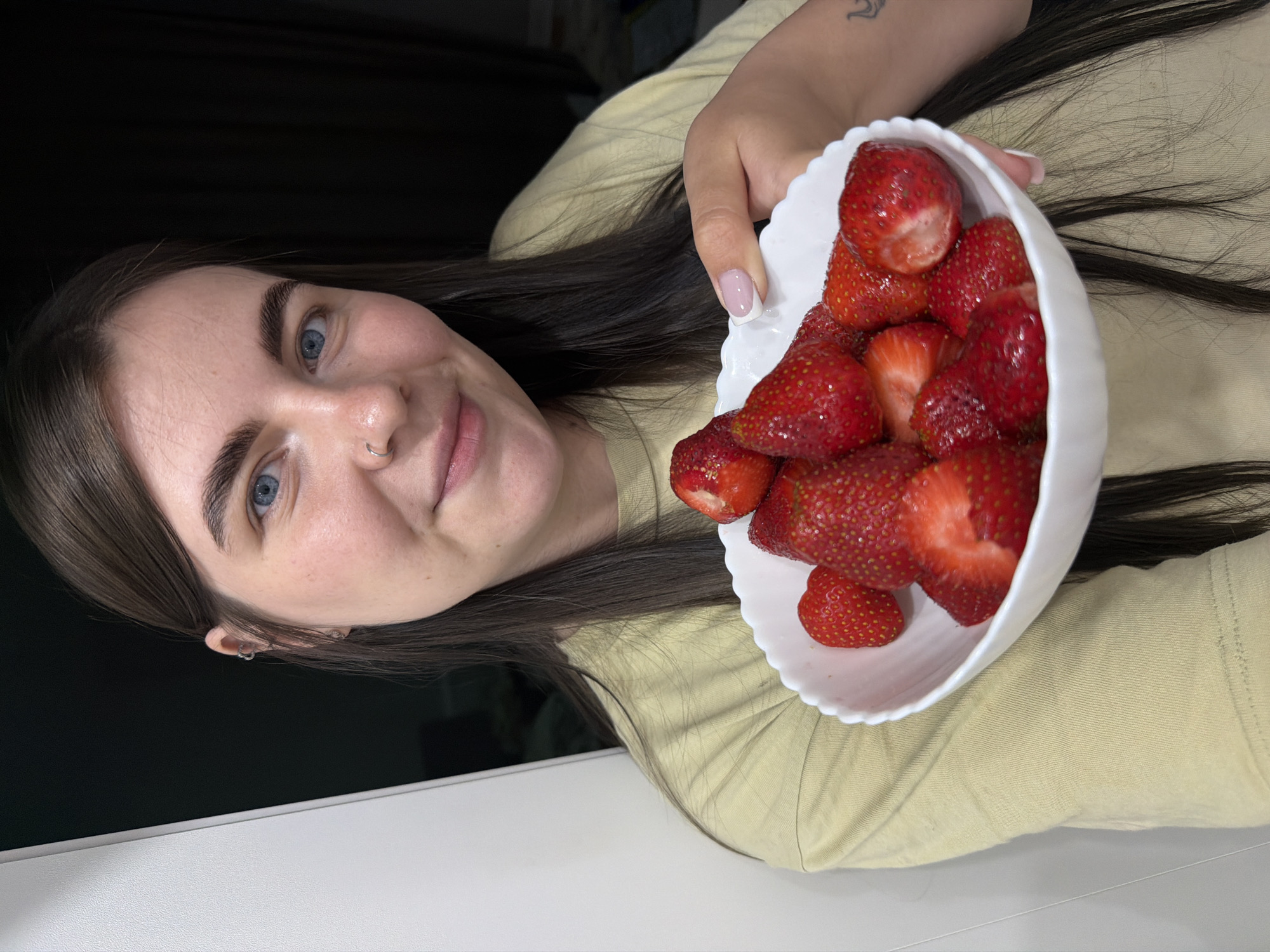A young woman with fair skin, blue eyes, and dark hair is holding a white bowl filled with red strawberries. She is smiling at the camera. She is wearing a light green shirt and has a nose piercing in her left nostril. Her fingernails are painted with a light pink color and white tips. Her hair is long and straight.
The background is dark. A white countertop is visible in the lower left corner of the image. The location is likely indoors. The city is not clear, or other details to confidently suggest that the image was taken in Chaikovsky, Russia, or any specific locale.