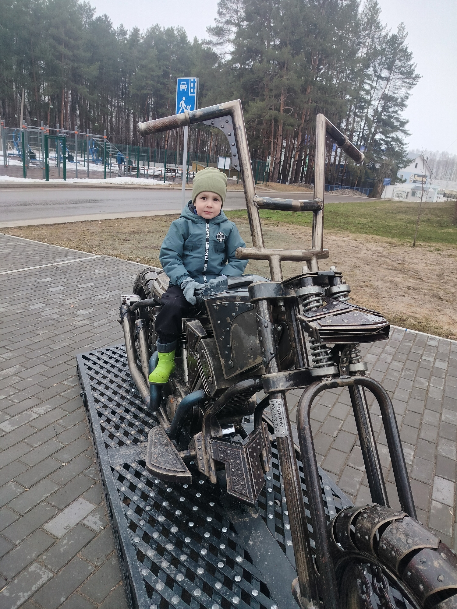A young child is sitting on a custom-built motorcycle sculpture in Chaikovsky, Russia. The child is wearing a green hat, a blue jacket, black pants, gray gloves, and bright green boots. The motorcycle sculpture is made of metal and rests on a metal grate platform that is situated on a paved area of gray bricks. In the background, there is a road, a park with exercise equipment, and a dense forest under an overcast sky. A blue sign with a pedestrian and wheelchair crossing symbol is visible in the background.