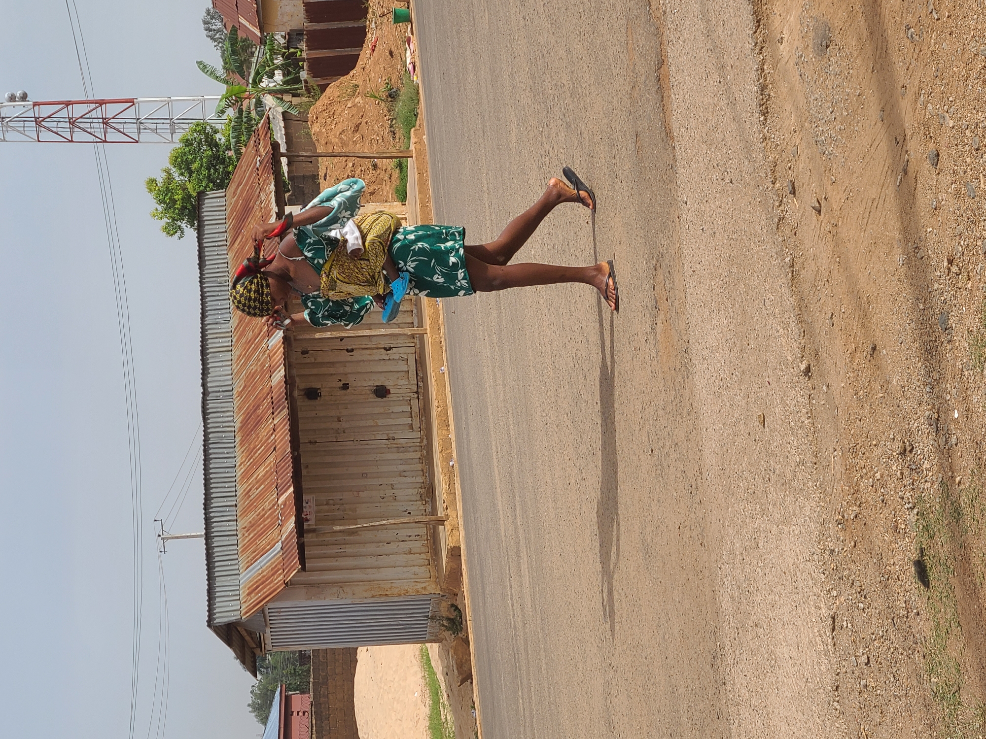 A woman in Gura Top, Nigeria, is walking on a road. She wears a headscarf and patterned clothing, and carries something wrapped in a cloth. A rusted corrugated metal building stands beside the road. In the background is a communication tower.