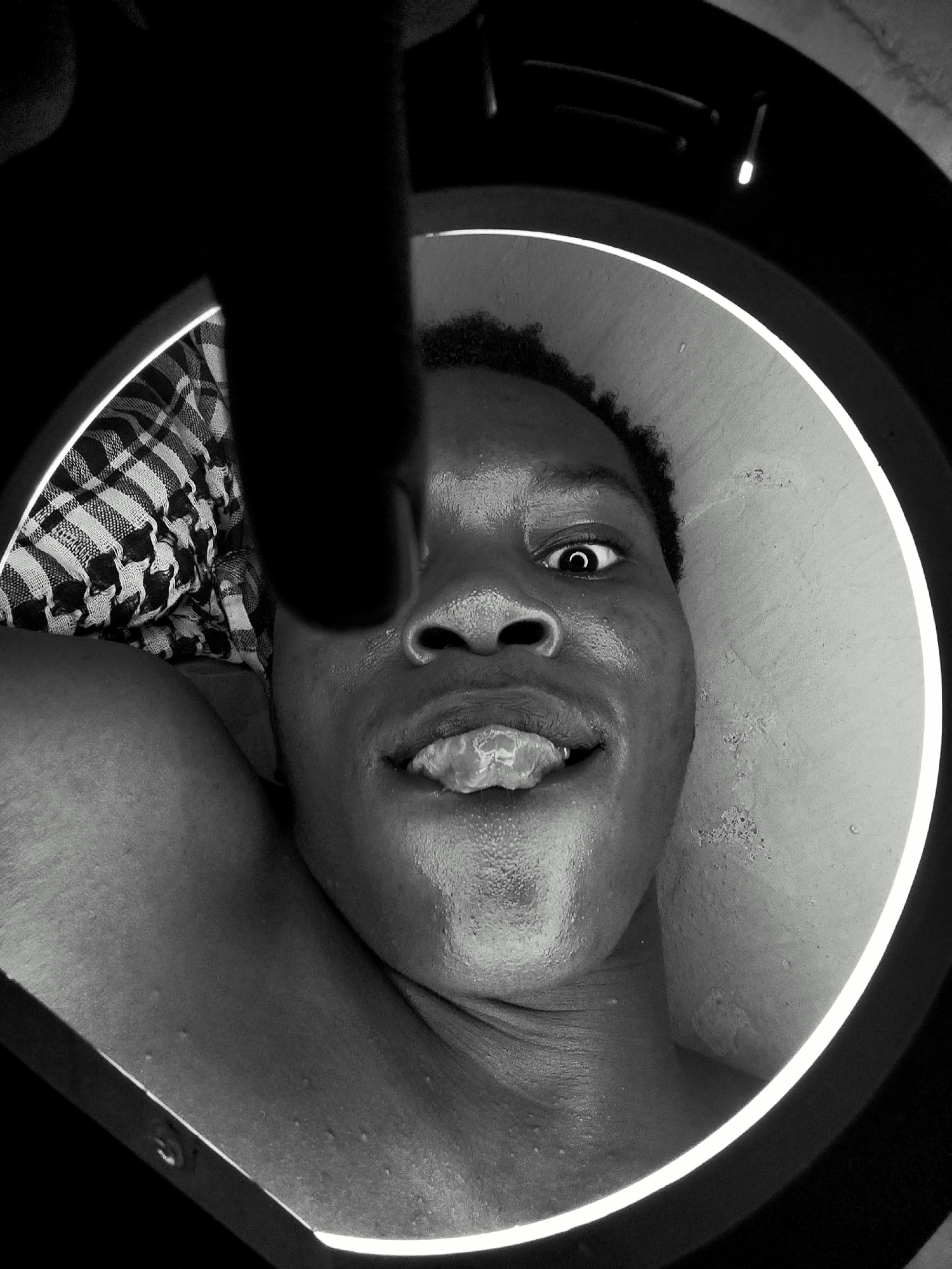A black and white close-up photo of a Black male's face taken from below, with his tongue sticking out, captured within the frame of a round object (possibly a ring light or similar). The background behind the subject is a light-colored surface, possibly a wall. Part of a patterned cloth is visible on the left, near the subject's shoulder. A dark shape is intruding into the frame from the top, obscuring a part of the subject’s face. The subject's right eye is wide and visible, with a pronounced expression. The location and city are unknown.
