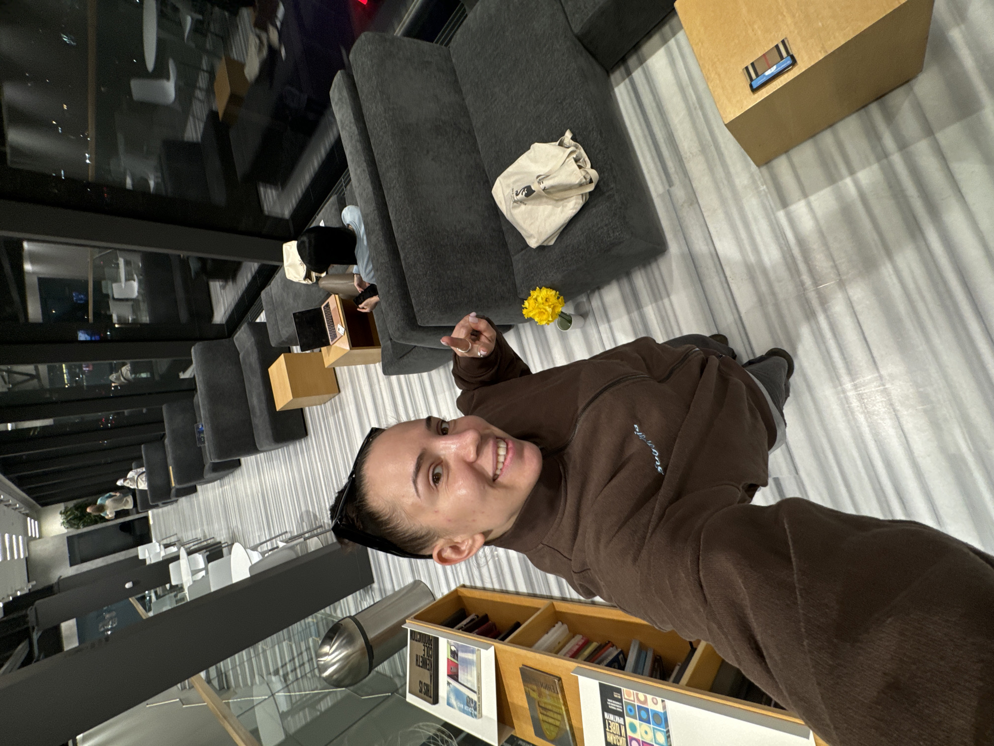 A woman in a brown turtleneck sweater is taking a selfie in a modern, well-lit indoor space. She's smiling and holding up one finger. The setting appears to be a contemporary lounge area with gray seating, a wooden side table, and a book display. A white tote bag sits on one of the couches, and a small bouquet of yellow flowers is placed nearby. Through large windows, a cityscape is visible, suggesting an urban location in Moscow, Russia. Other people are seated in the background, using laptops. The floor is striped with light and dark lines.