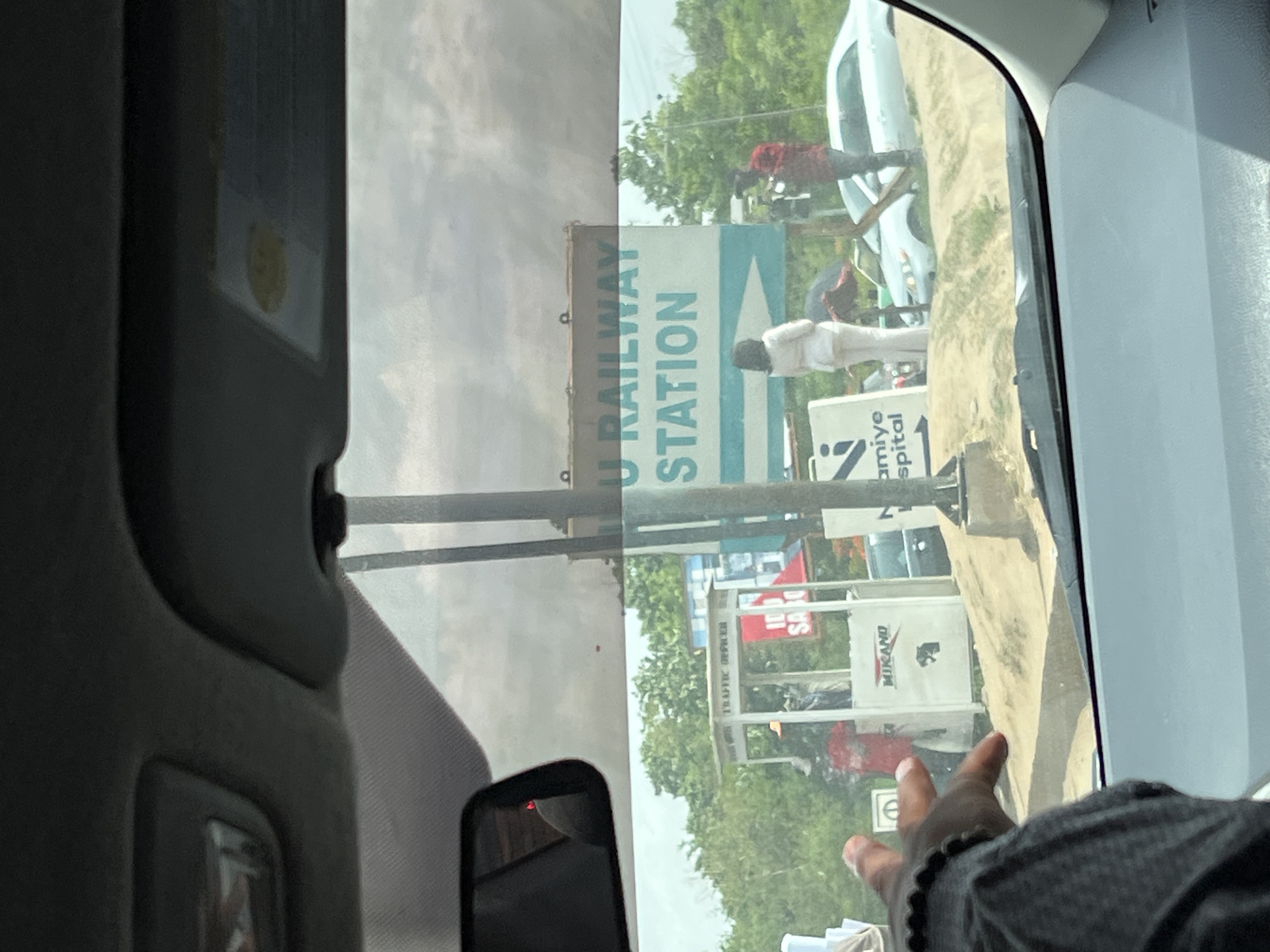 The image captures an outdoor street scene in Karmajiji, Nigeria, viewed from inside a vehicle under bright daylight. The perspective is through the passenger side window, with reflections and vehicle interior elements partially obscuring the view.
In the foreground, the dark interior of the car is visible, including part of the dashboard/visor with a small, yellow, possibly smiling face sticker. A smartphone rests in the lower-left corner, reflecting light. From the bottom-right, a hand wearing a beaded bracelet extends into the frame.
Outside the vehicle, several people are present. One individual, dressed in a white garment, stands near a prominent road sign. Another person in a red top and dark trousers is visible further back, near a white car.
Multiple signs line the roadside. A large, vertical sign with white text on a blue-green background reads "U RAILWAY STATION," likely indicating a railway station ahead. Below this, a smaller white sign with an arrow points to "amiye Hospital" (possibly part of "Karamiye Hospital"). Further in the background, a red billboard partially shows "100 DAYS" and "SA," with a small sign underneath reading "TRAFFIC OFFICER." Another white sign displays "MIKANO" and a dark animal logo, possibly an elephant or rhino. The background features green trees and areas of dry, uneven ground. The overall scene suggests a routine day in an urban or semi-urban area, with people and vehicles by the roadside.