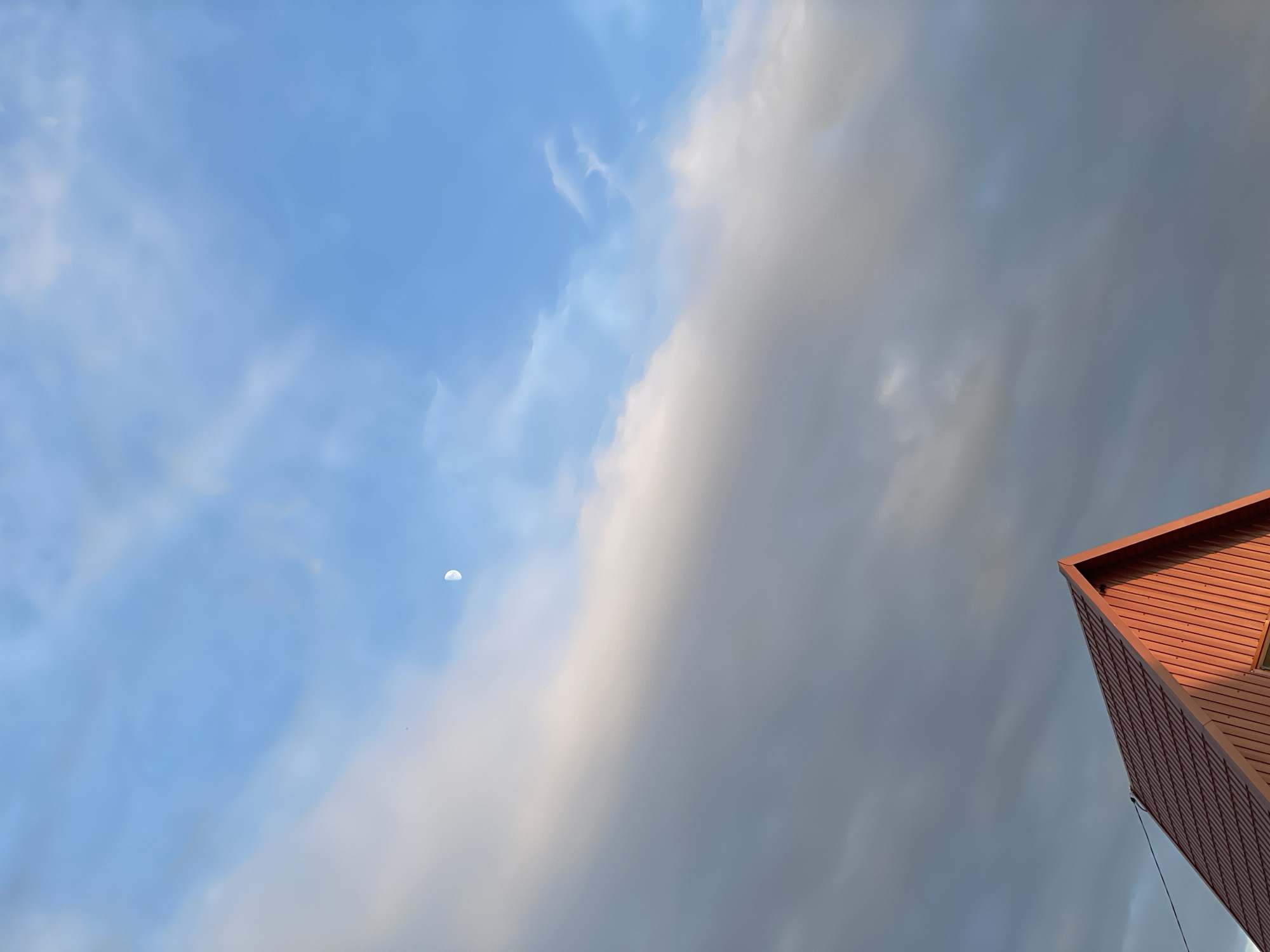 The image captures a dramatic sky scene with a crescent moon visible in the upper left quadrant. The sky is divided between a bright, clear blue on the left, dotted with wispy white clouds, and a darker, more overcast expanse of grey clouds on the right. The division creates a striking contrast. In the lower right corner of the image, the sharp, angular edge of a modern building with reddish-brown siding is visible, reaching towards the sky. The building's architecture is geometric and contemporary. The lighting suggests either early morning or late afternoon, as the sun casts a warm glow on the side of the building, while the sky exhibits a mix of daylight and approaching or receding clouds. There is no visible text in the image. The overall impression is one of a vast, dynamic sky meeting the man-made structure, creating a sense of scale and perhaps a hint of atmospheric grandeur. The weather appears to be changing, with a mix of sunshine and cloud cover. The location is identified as Buynaksk, Russia.