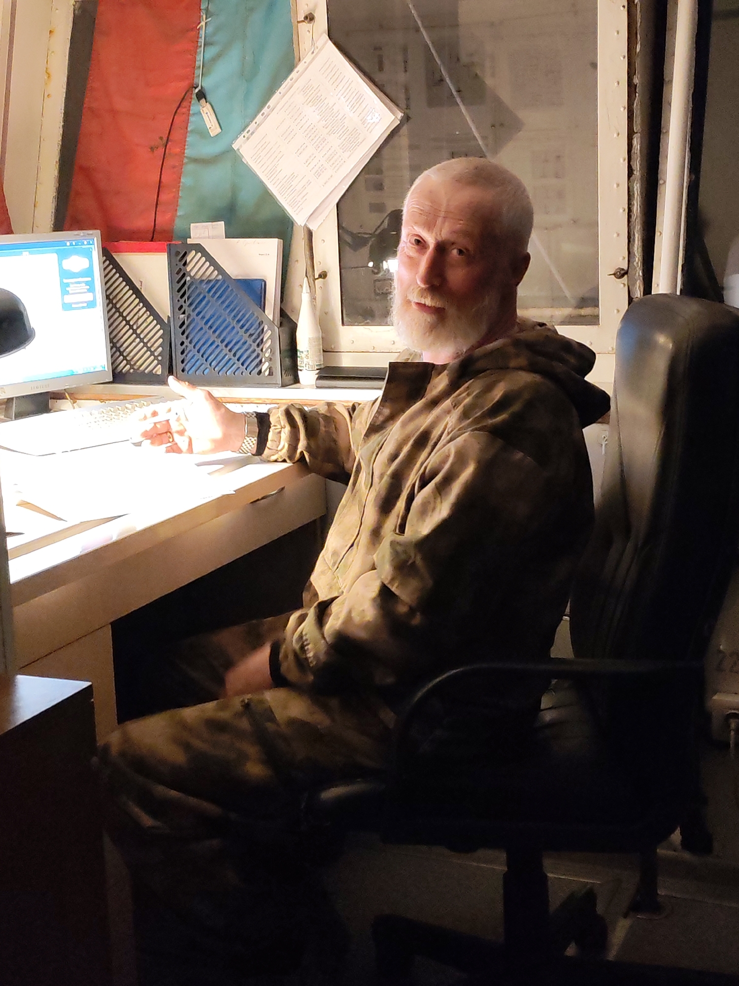 An adult male with a white beard and short white hair sits at a desk in what appears to be an office or control room. He is wearing a camouflage jacket. The desk is white, and there is a computer monitor and keyboard on the desk, along with some papers and file organizers. Behind the desk are red and blue flags. There is a window behind the man. The man is seated in a dark office chair. The location is possibly in Yakhroma, Russia.