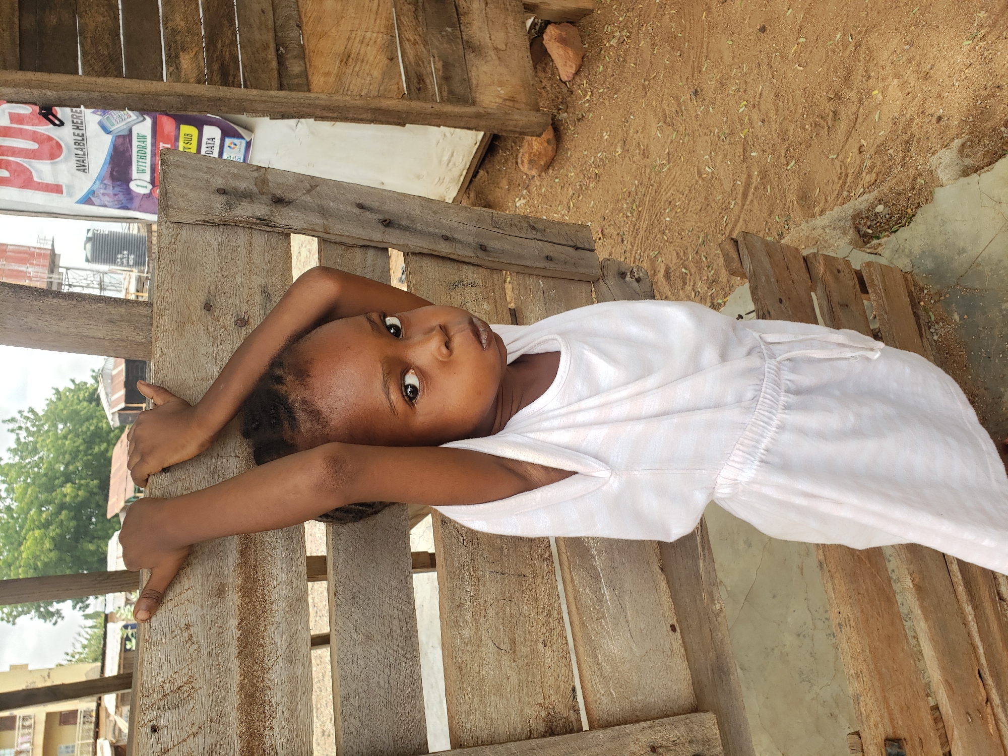A young child, appearing to be a girl, lies on her back on a rustic wooden structure, looking directly at the camera. Her arms are stretched out and rest on the rough wooden planks of the surface she is on. She wears a simple, light-colored dress or top. Her expression is calm and direct, with a hint of curiosity in her wide eyes.
The setting appears to be outdoors or within a simple, open structure. The ground around the wooden platform is sandy and reddish-brown, scattered with small leaves or debris. In the upper left background, a colorful banner or sign is partially visible. Text on the banner includes "AVAILABLE HERE", "WITHDRAW", "TV SUB", and "DATA", suggesting various services offered, possibly related to mobile banking or telecommunications. Part of a larger word, possibly "PAY" or "PU", is also visible on the banner.
Beyond the immediate foreground, distant buildings with a black water tank on a rooftop and some green foliage are visible, suggesting an urban or semi-urban environment. The lighting indicates it is daytime. The scene captures a candid moment in Adankolo, Nigeria.