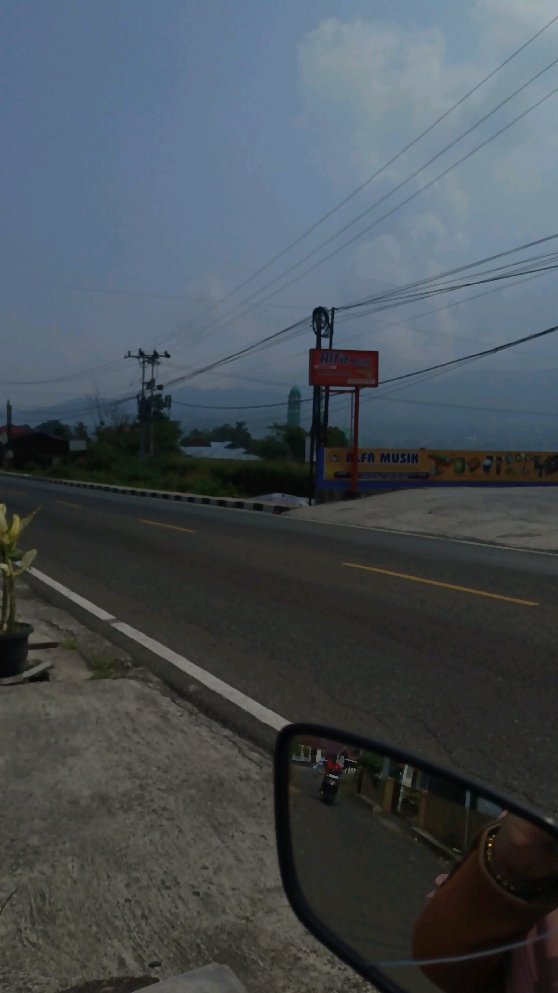 The image depicts a roadside scene with a paved road and sidewalk. A motorcycle is reflected in the rearview mirror on the right side of the frame, with its rider wearing a red shirt. Further down the road, a billboard with "ALFA MUSIK" is visible, along with an orange sign. In the background, power lines crisscross the sky, and a tall, slender building, possibly a mosque minaret, stands to the left of the billboard. The landscape is generally hilly with some vegetation. The sky is overcast with clouds, suggesting it might be dawn or dusk. The location is Panyalaian, Indonesia.