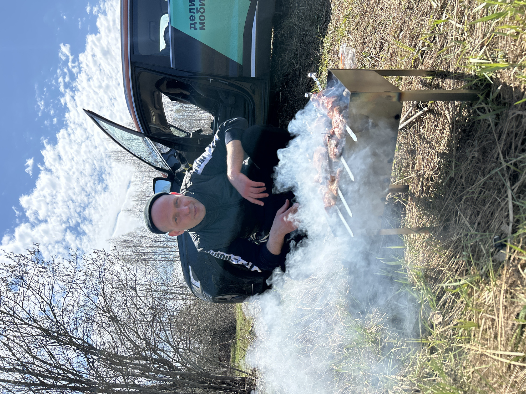 A man in a black jacket and a black hat is kneeling in front of a barbecue grill. The grill is smoking, and there are skewers of meat on it. The man is looking at the camera. Behind him, there is a car with an open door. The scene is set in a grassy area with trees in the background. The sky is blue with some clouds. The location is Unknown, Unknown.