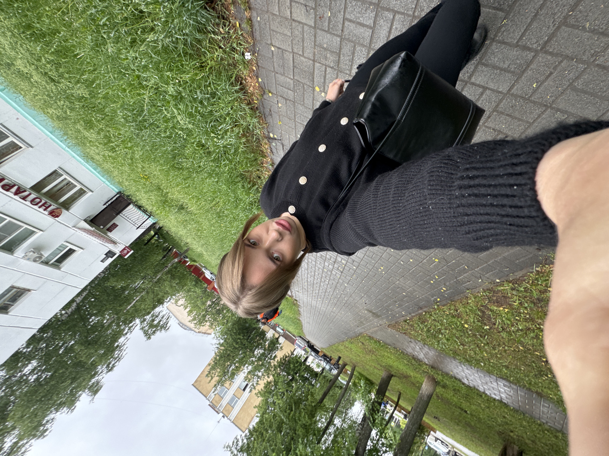A woman with blonde hair is taking a selfie outdoors. She is wearing a black sweater with gold buttons and black pants. A black purse is visible over her shoulder. She is standing on a paved sidewalk next to a patch of green grass. In the background, there is a building with the Cyrillic word "НОТАРИУС" (Notary) visible on its facade, indicating the city of Chaikovsky, Russia. The weather appears to be overcast. Other buildings and trees are also in the background.