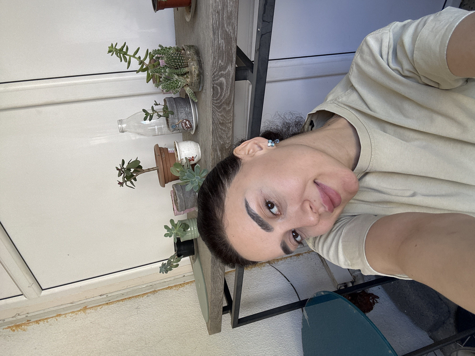 A person is visible in a medium shot. The person has dark hair pulled back, fair skin, and is wearing a light-colored t-shirt. The person is looking at the camera with a slight smile. Behind the person is a wooden table with several potted plants. The plants appear to be succulents. The background is a white wall with a window. The location is likely in Tbilisi, Georgia, based on the presence of succulents, which are commonly grown in this region. A blue chair is also visible.
