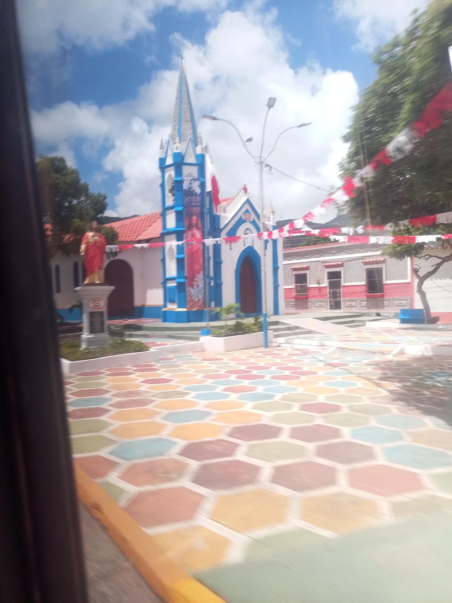 The image captures a street scene in Tacarigua, Venezuela, focusing on a vibrant plaza in front of a church. The foreground is dominated by a colorful hexagonal pavement, with tiles in shades of yellow, orange, pink, green, and blue. To the left, a dark, blurry frame, likely from a vehicle window, partially obscures the view.
In the mid-ground, a white church with blue trim and a tall, pointed steeple stands prominently. A statue of a figure in a red robe, possibly religious, is positioned on a pedestal to the left of the church entrance. The church is decorated with blue and white banners featuring images of religious figures. Adjacent to the church, a building with a red tiled roof and pink and gray walls features arched windows. Red and white festive banners stretch across the upper portion of the scene, strung between lampposts and trees.
The background shows lush green trees and a mountainous landscape under a sky filled with white clouds. The overall impression is one of a bright, lively public space, likely decorated for a celebration or event. The image appears to be taken from a moving vehicle due to the motion blur.