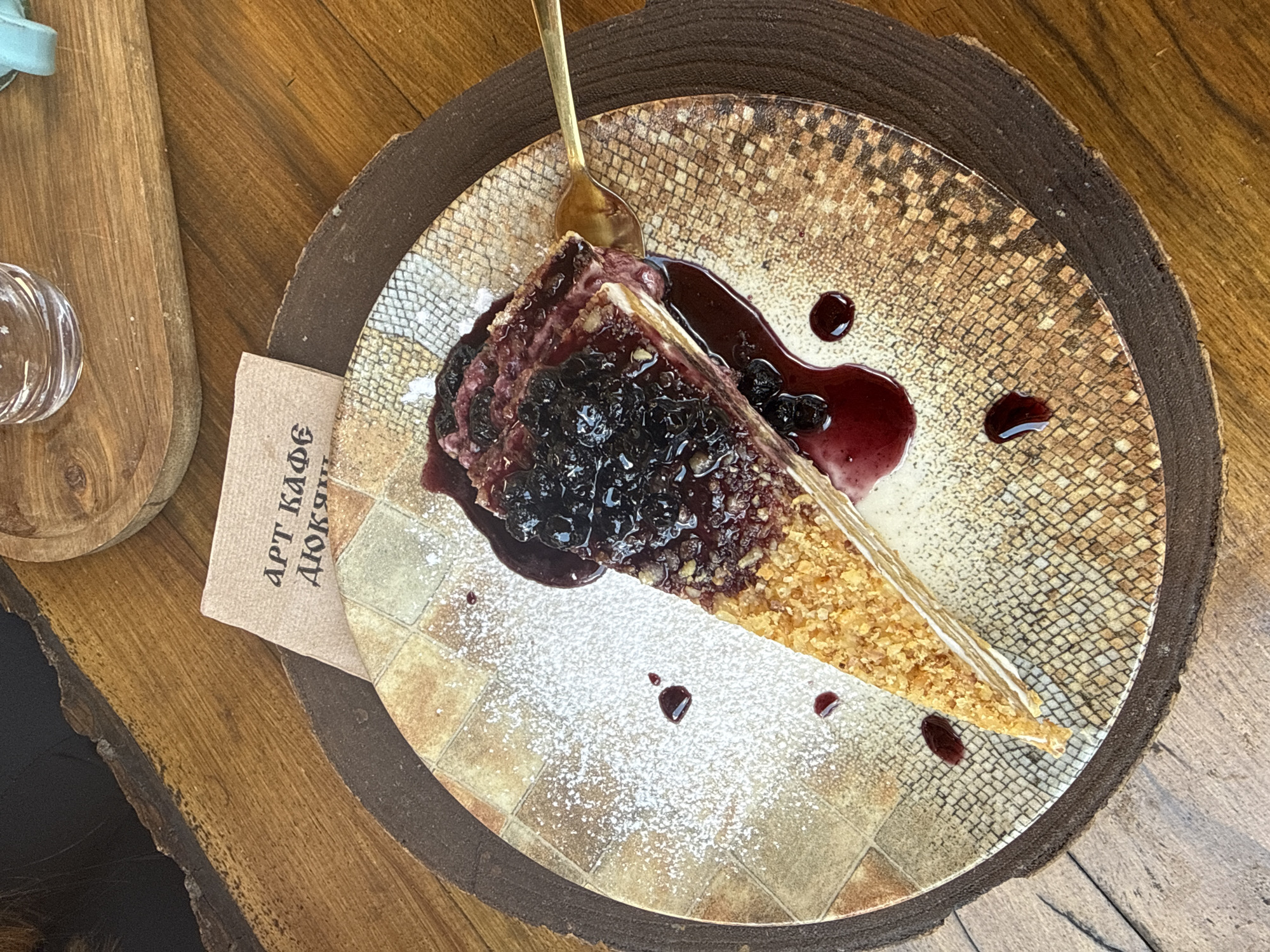 A slice of layered cake, topped with a dark berry sauce and fresh berries, sits on a patterned plate on a wooden table. A gold fork is inserted into the cake. A small brown card with Cyrillic text is placed behind the plate, and a glass is visible in the background on another wooden surface. The scene is located in the city of Shiroka Laka, Bulgaria.