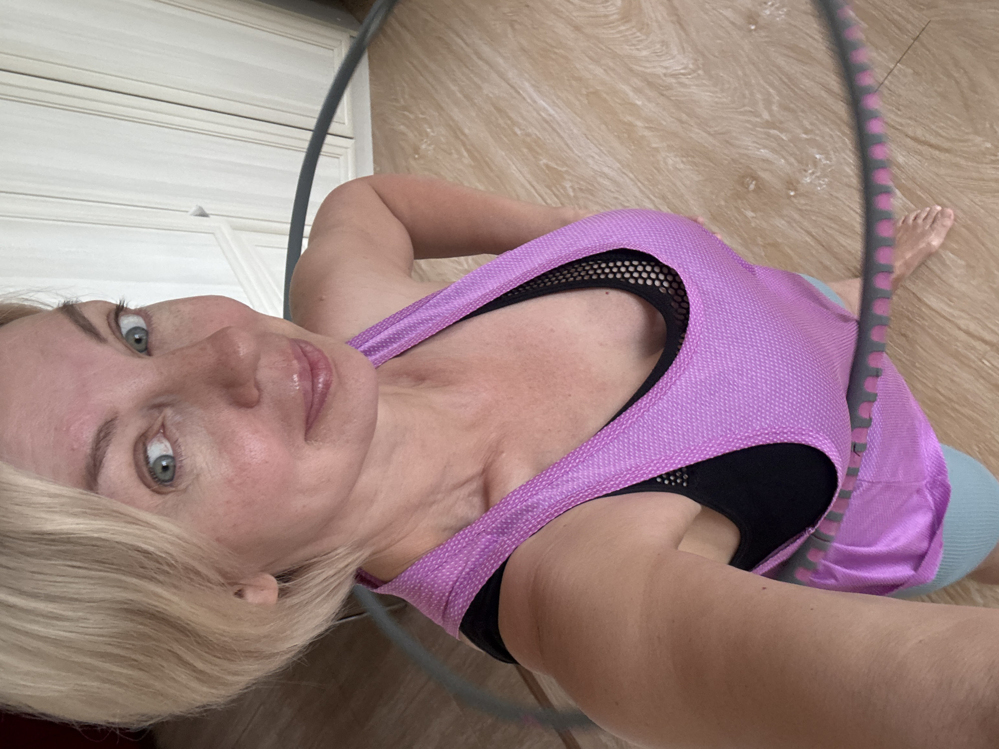 A woman with blonde hair is posing for a selfie while holding a hula hoop. She is wearing a pink tank top over a black sports bra and light blue shorts. The background shows a wooden floor and a light-colored cabinet. The image was taken indoors, likely in a residential setting. There is no indication of the location being in Sochi, Russia.