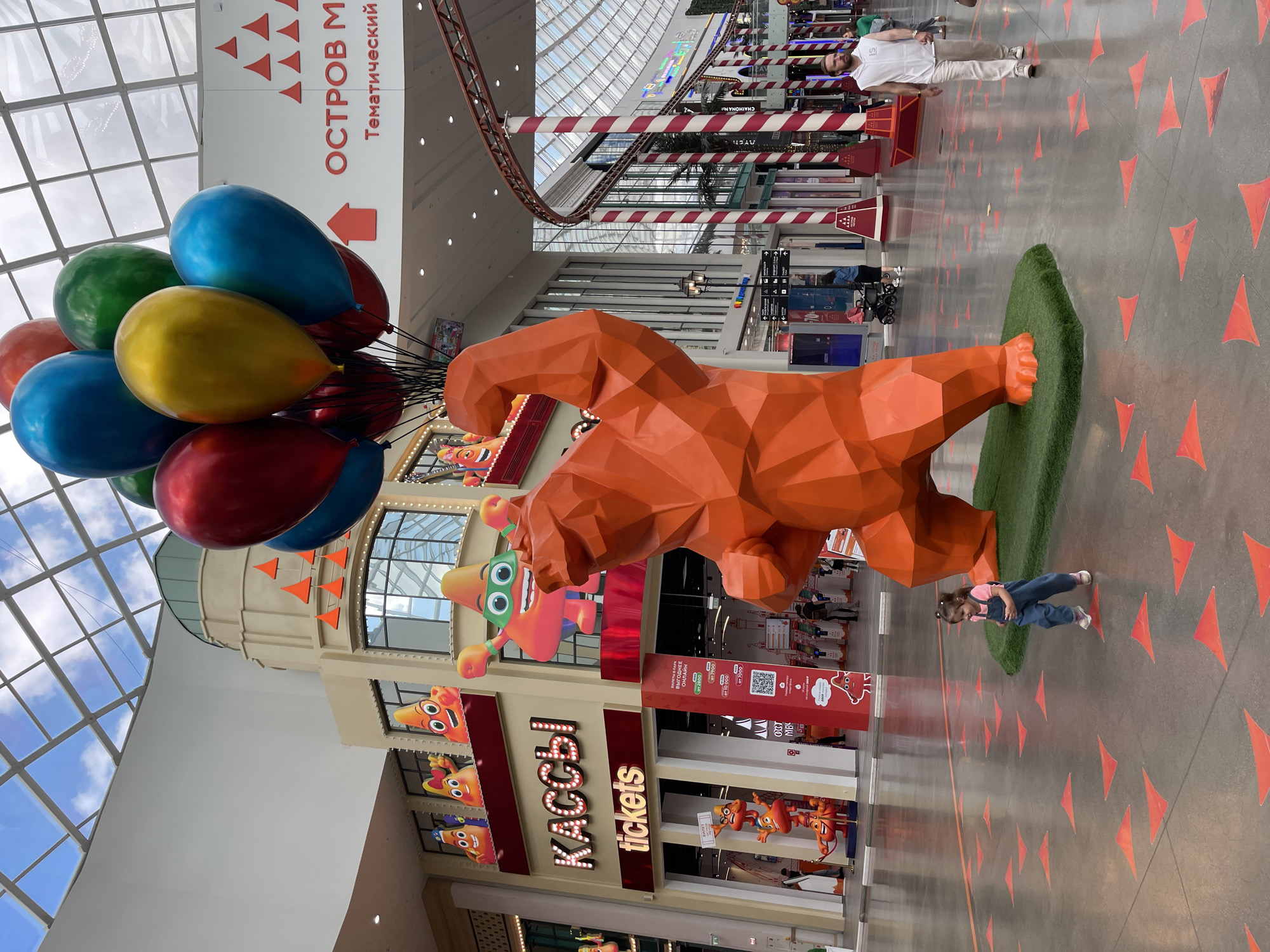 An orange, polygonal bear sculpture is the central focus of the image, standing on a patch of artificial grass in an indoor public space. The bear is depicted with its left arm raised, flexing its bicep, and is holding a cluster of large, colorful balloons (red, blue, yellow, and green) with black strings. To the left of the bear, a young child wearing a pink shirt and blue overalls is walking on the polished concrete floor, which is decorated with scattered red triangle-shaped markers. In the background, a building facade with illuminated "КАССЫ" (meaning "TICKETS") signage is visible, along with cartoonish orange cone-shaped characters. Further back, a roller coaster track winds through the space, and various shops and people can be seen. The ceiling is a glass-paned structure, allowing natural light to filter in. A man in a white sleeveless shirt and light-colored pants walks past the bear from right to left.