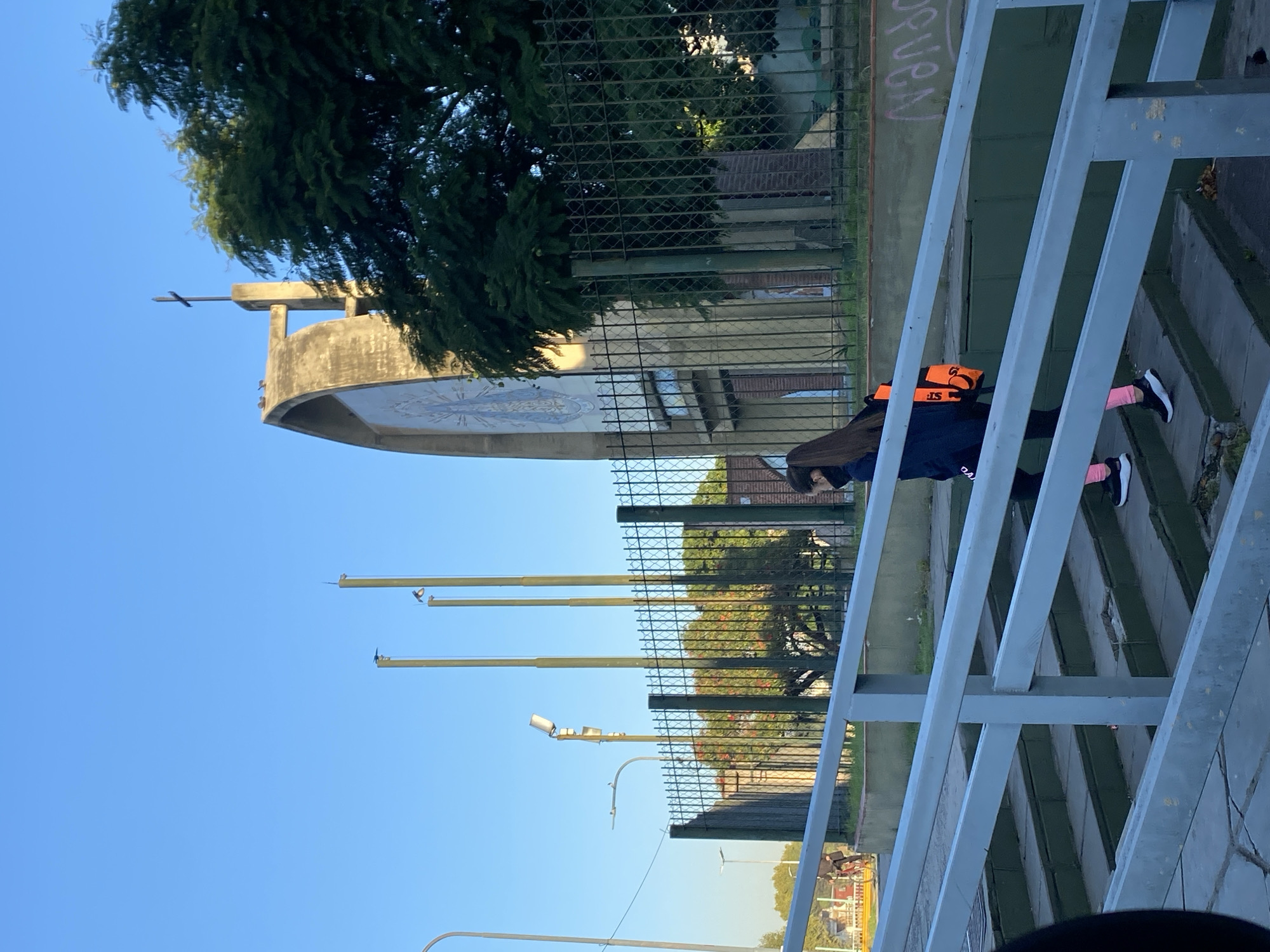 A partially obscured building with a cross atop is visible behind a chain-link fence. In front of the fence, a person is walking. This scene likely depicts the location near the sanctuary of Our Lady of Schoenstatt in the city of Buenos Aires, Argentina.