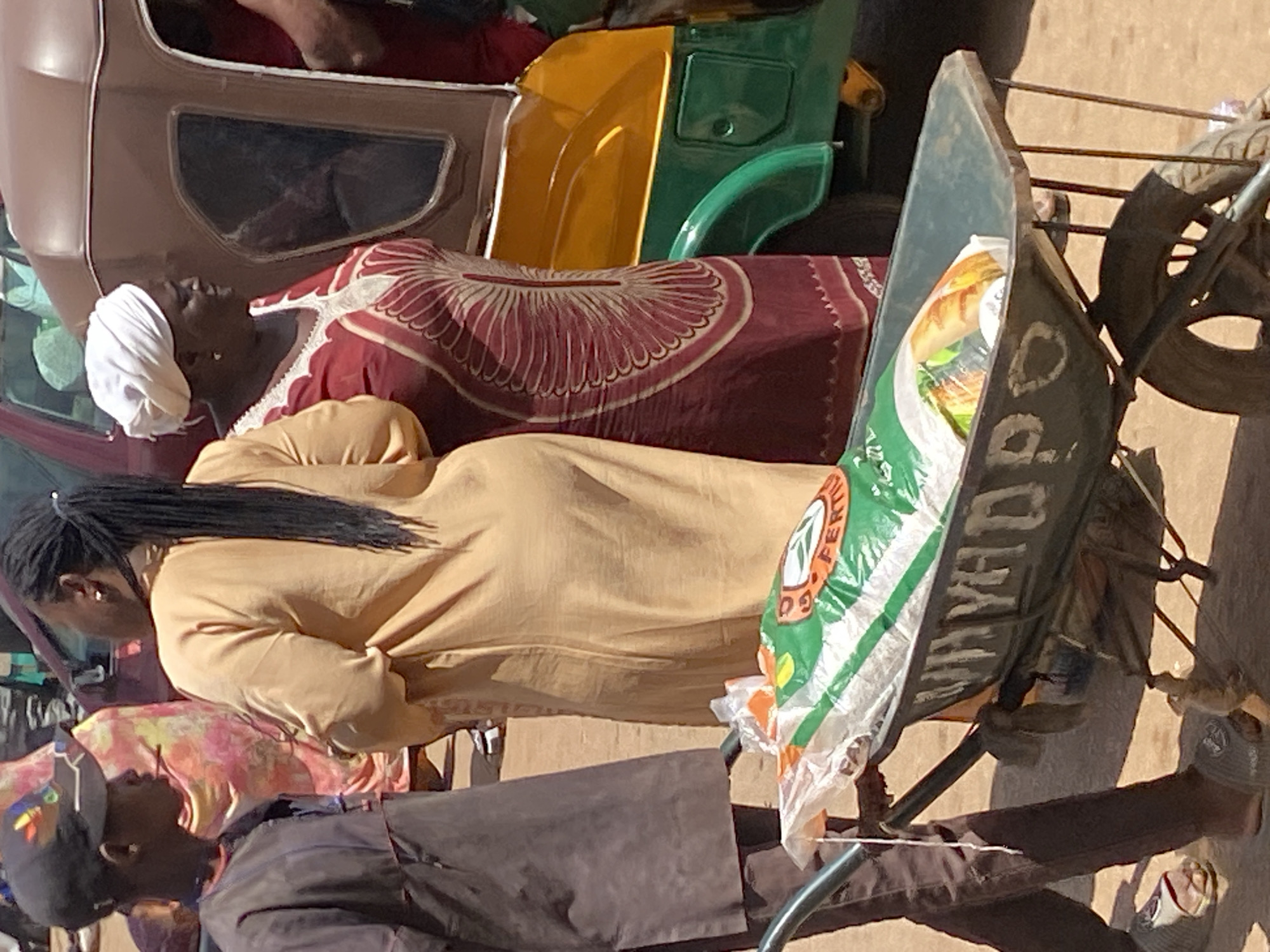 This image captures an outdoor scene in Bukuru, Nigeria, during daylight hours, featuring several individuals and vehicles, suggesting a bustling public or commercial area. The ground appears to be a light, sandy color.
In the foreground, a woman with dark braided hair, wearing a light brown or beige long-sleeved top, is standing or leaning, facing towards the left side of the frame. Below her, a man with dark skin, wearing a dark cap and dark jacket, is partially visible, looking upwards.
Prominently featured is an auto-rickshaw, commonly known as a "keke napep" in Nigeria. Its body shows sections of brown, yellow, and green. Adjacent to the auto-rickshaw, a woman with dark skin and a white head covering is visible, appearing to be seated or resting in the vehicle's opening. She is wearing a maroon-colored garment with an intricate white and red circular pattern.
To the right, a dark green or black metal wheelbarrow or cart is positioned. On its side, the word "MAYAD" is visible. Inside the cart, a large white and green bag is placed. Partially visible text on this bag includes "FERTI GO" and "FERTILIZER," along with a leafy logo, suggesting its contents might be agricultural or food-related. What appears to be a long loaf of bread or baguette is peeking out from the top of this bag. Further right, a bicycle or motorcycle wheel with spokes is visible. The bare foot of a person can be seen in the upper right, possibly from inside the keke napep, and another person wearing sandals and dark trousers is partially visible in the lower right, near the wheelbarrow.
The activity suggests people are either commuting, waiting, or engaging in commerce or transport of goods within the described setting. The clear lighting indicates it is daytime with good visibility.