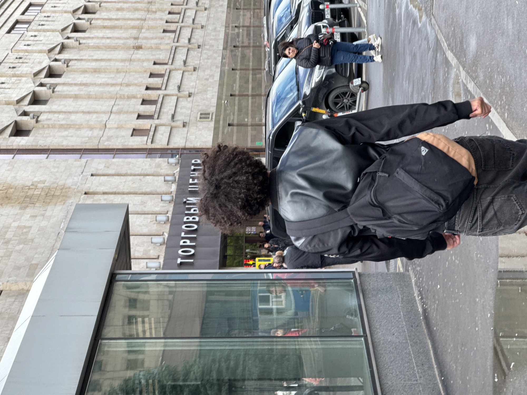 The image depicts an urban scene in Moscow, Russia, showing a sidewalk adjacent to a building with signage reading "ТОРГОВЫЙ ЦЕНТР" (Shopping Center). A person with curly hair, wearing a black jacket, jeans, and a backpack, is standing on the sidewalk. Behind this person, parked cars are visible. Another person in a black jacket and blue jeans is walking near the cars. The background includes the facade of a building with a distinctive architectural style. The ground appears wet, suggesting recent rain.