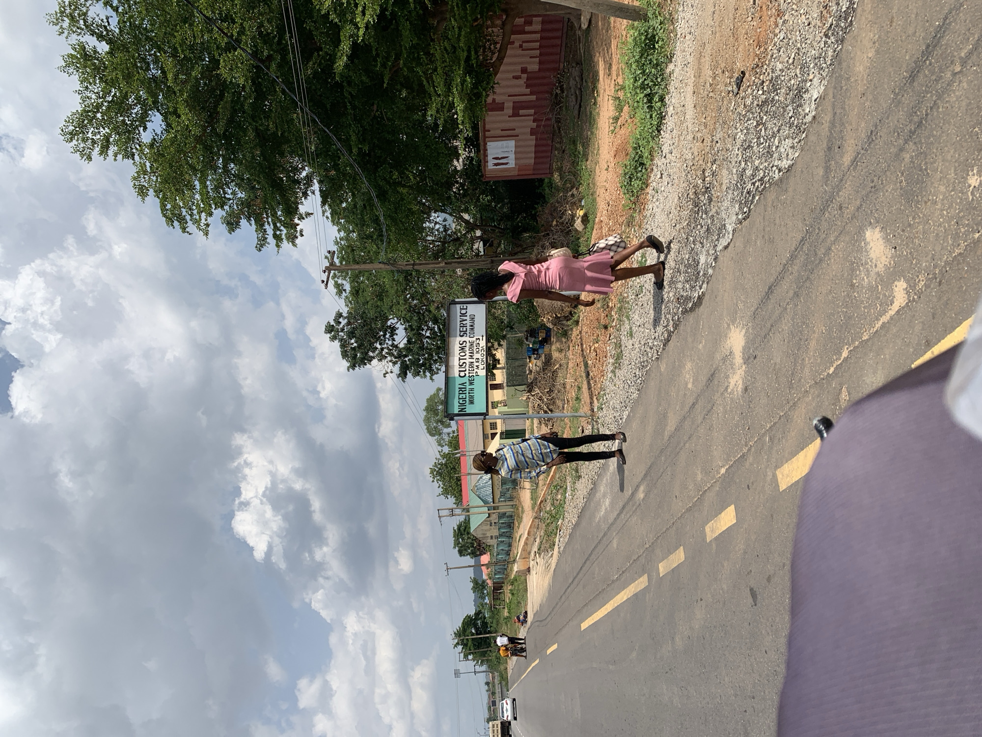 The image shows a road in Lokoja, Nigeria, with a sign for the Nigeria Customs Service North Western Marine Command. Two individuals are seen on the side of the road; one wearing a pink dress and the other a blue and white striped shirt. Yellow lines are painted on the road. Trees and buildings are visible in the background under a cloudy sky. A vehicle is also present in the distance.