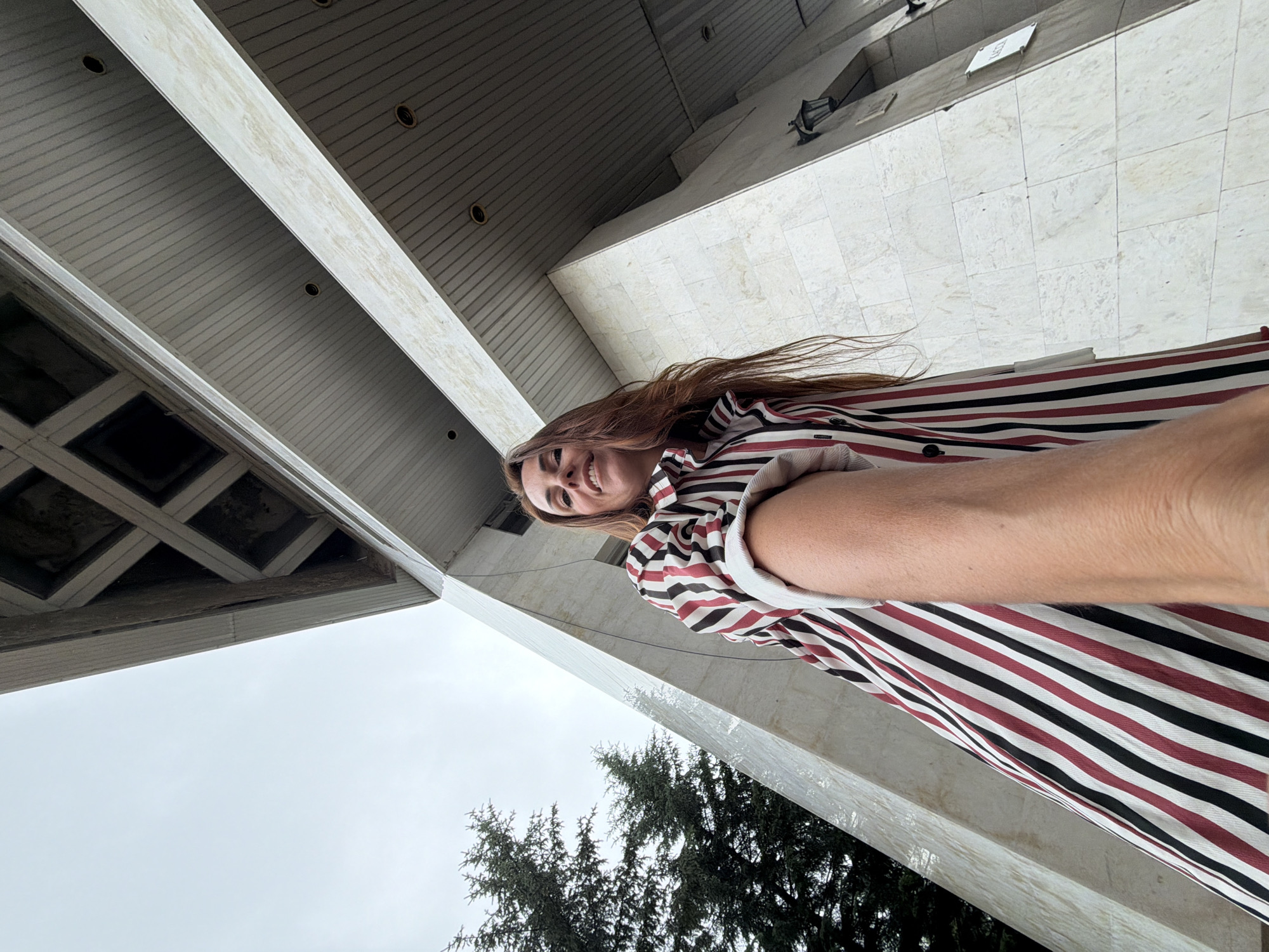 A low-angle outdoor, full-body shot shows a woman in a striped shirt standing in front of a large, modern concrete building with a cloudy sky above. The woman has long, reddish-brown hair and is smiling at the camera. She is wearing a collared shirt with vertical stripes in red, black, and white. Her right arm is extended towards the camera, and her left arm is bent at the elbow. The building is angular and imposing, with large concrete panels and a textured ceiling. A few trees are visible to the left of the building. The overall impression is of a modern urban environment.
