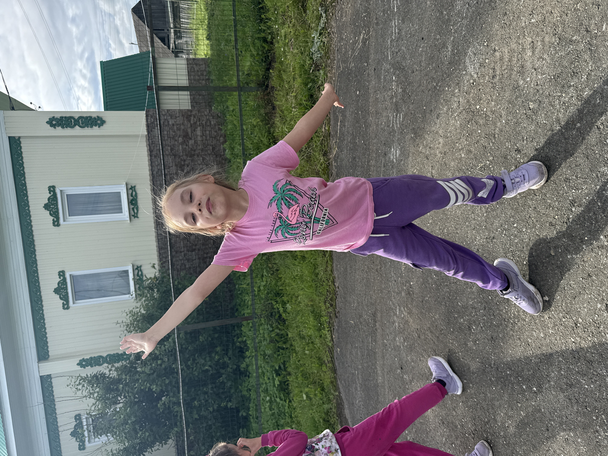 In Bolshoy Turysh, Russia, a young girl with blonde hair and light skin is standing outdoors on a paved surface, facing the camera. She is wearing a pink t-shirt with a graphic design and purple sweatpants. Her right arm is raised, with her hand open. To her left, another child, partially visible, is wearing pink sweatpants. The background shows a light-colored building with green trim, a green fence, and lush green vegetation. The sky above is partly cloudy. The overall scene appears to be a residential area on a daytime, possibly summer, day.