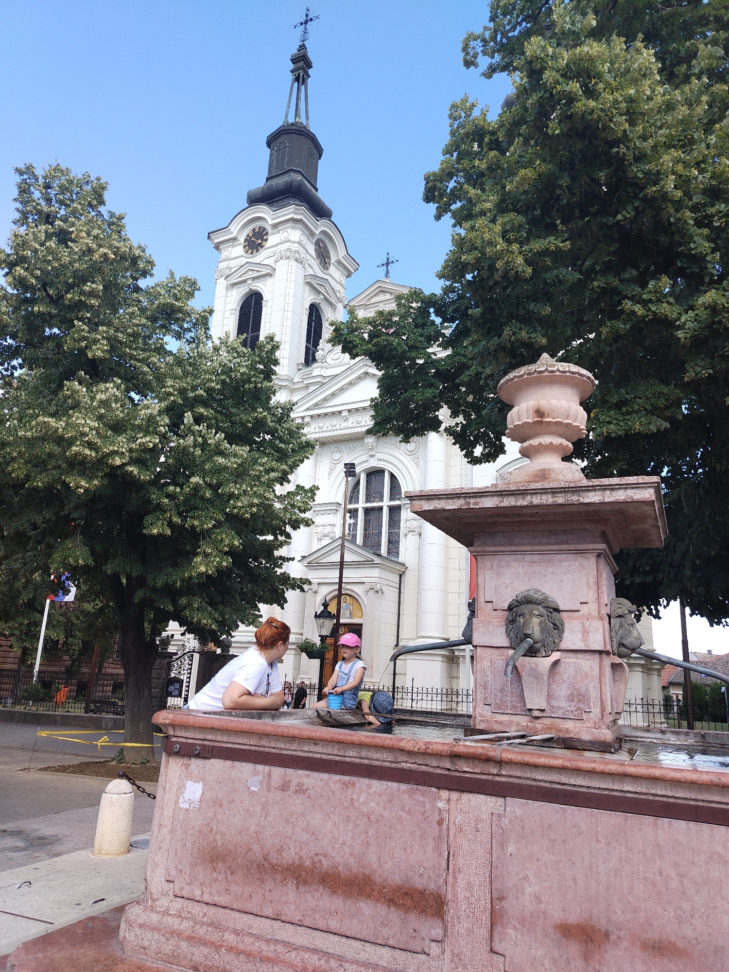 In the city of Sremski Karlovci, Serbia, a woman with red hair and a white t-shirt leans on the edge of a large, reddish-brown stone fountain. She is looking down at a young child wearing a pink hat and denim overalls, who is sitting on the fountain's edge and playing with a blue bucket. The fountain features two lion heads with spouts. Behind the fountain, a large, white church with a tall clock tower and a cross on top is visible. Lush green trees frame the church and the scene. The sky is clear and blue.