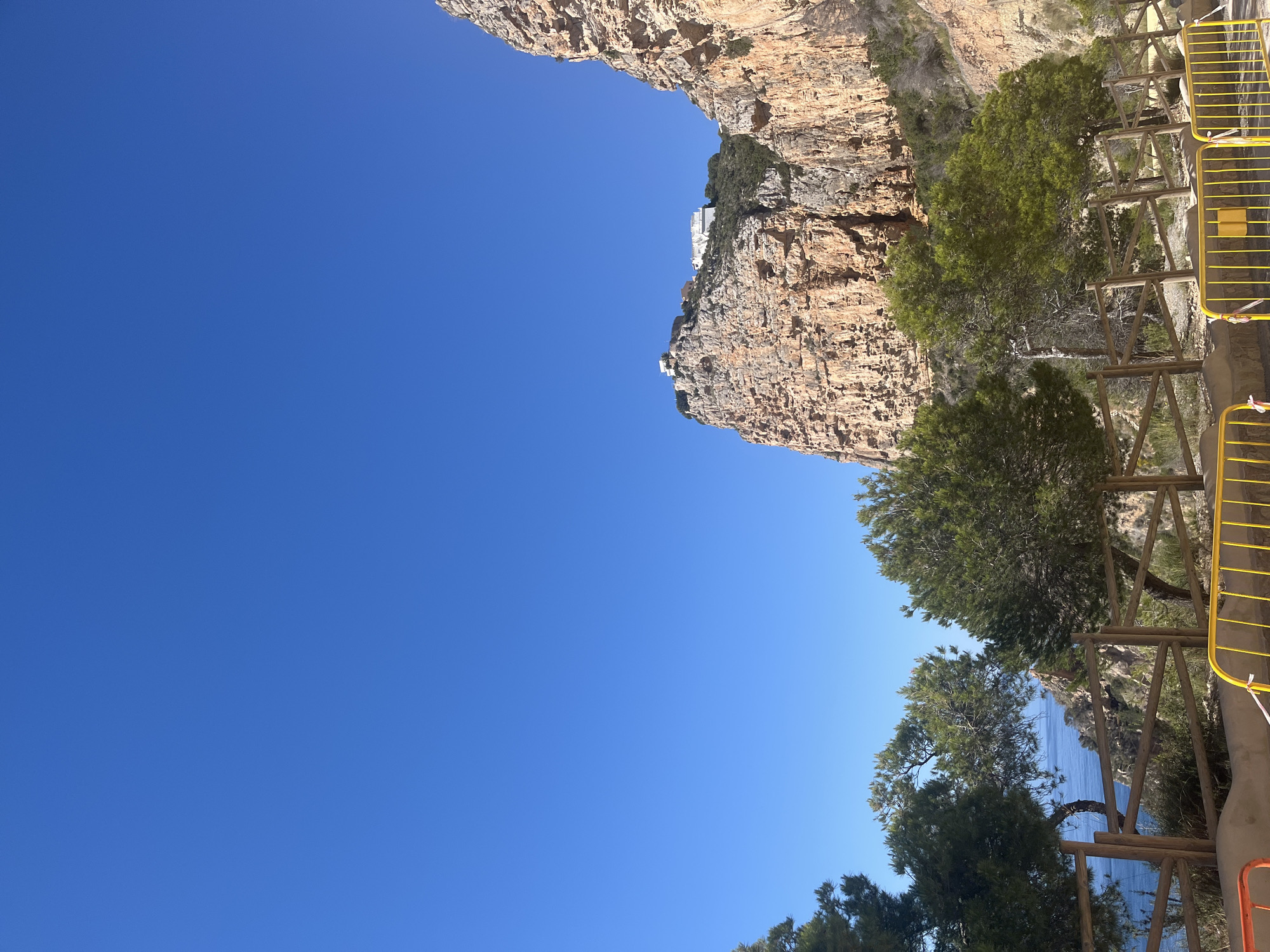 The image depicts a view in el Poble Nou de Benitatxell / Benitachell, Spain, showing a rugged, orange-hued cliff face descending towards the sea, with a bright blue sky dominating the scene. Vegetation is present, including trees and shrubs growing along the cliff's edge and near a wooden fence with yellow metal railing. A small white building is perched atop the cliff, accentuating the dramatic landscape. The sea is visible at the lower edge, suggesting a coastal setting.