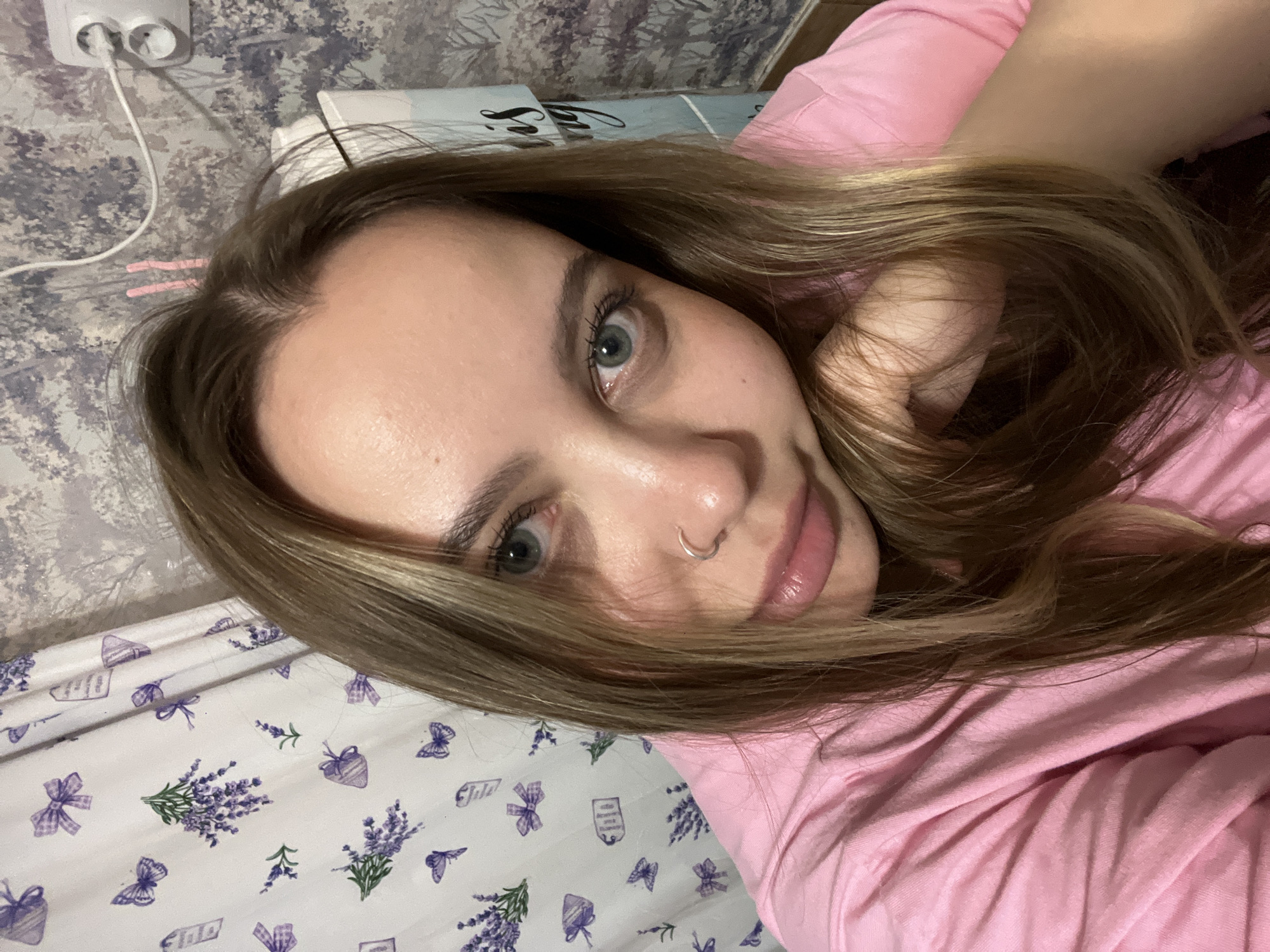 The original image displays a portrait-oriented selfie of a young woman taken indoors, likely in a bedroom in Novopokrovskaya, Russia. The woman, who appears to be in her late teens or early twenties, lies on her side, looking directly at the camera. She has fair skin, light brown or ash-blonde hair that frames her face, and striking blue-green eyes. A silver ring nose piercing is visible on her right nostril, and her lips are slightly parted with a glossy finish. Her expression is calm and neutral, with a soft gaze.
She is wearing a casual pink top, and her right hand is partially visible within her hair on the right side of the frame, possibly supporting her head. The background on the upper left features a wall with a subtle, patterned wallpaper. A white electrical outlet with two white charging cables plugged in is mounted on this wall. Small pink hair clips are discernable near her hairline on the upper left. The lower left portion of the image reveals bedding, specifically a white sheet or pillowcase, decorated with a repeating pattern of purple lavender sprigs, ribbons, and butterflies, accented with green leaves. A white, possibly rectangular object, such as a book or another pillow, is partially visible behind her head. The lighting is artificial and soft, suggesting an indoor setting without strong natural light. No discernible text is visible within the frame.