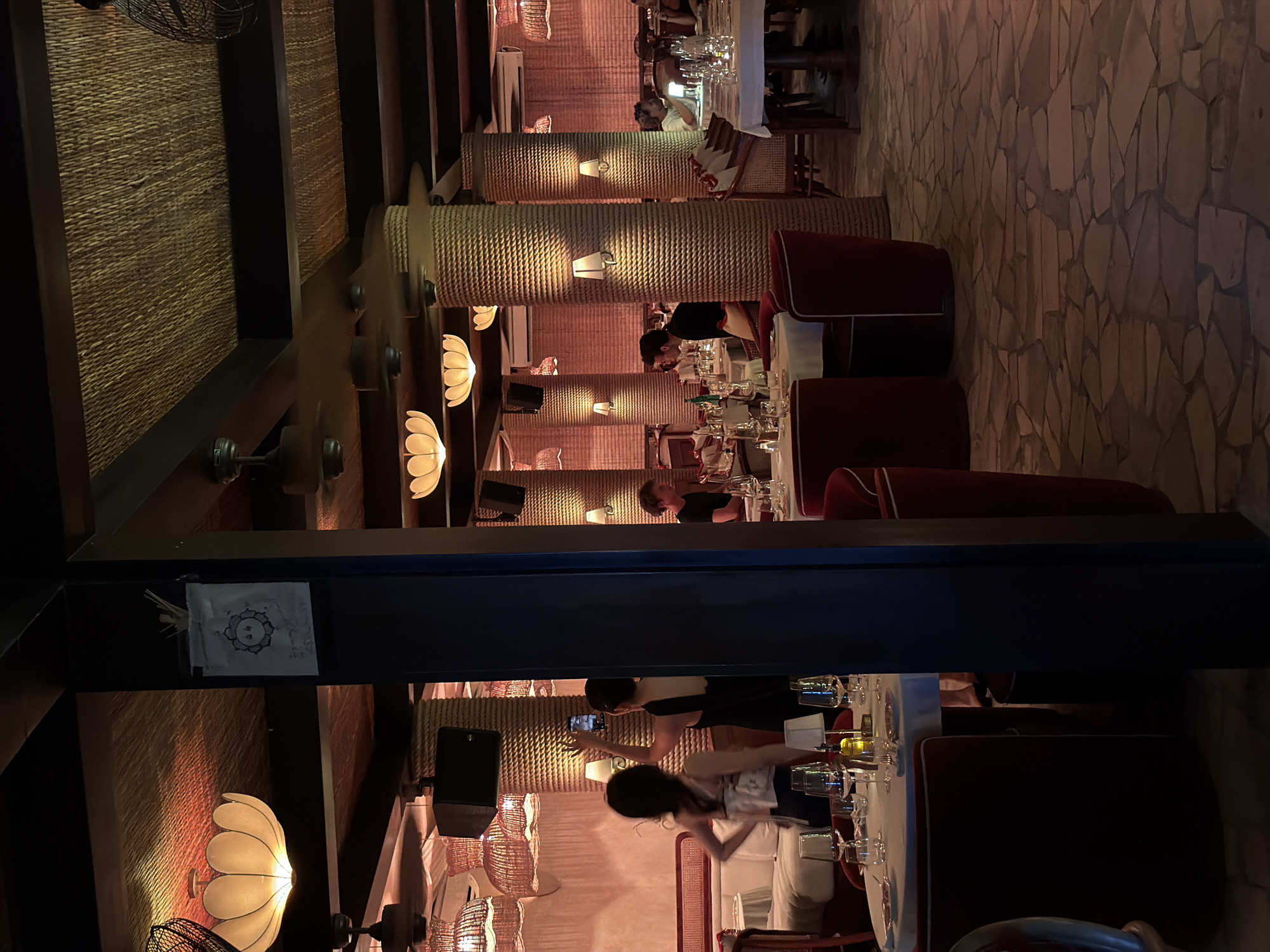 The image shows the interior of a restaurant in North Kuta, Indonesia. The restaurant has a rustic and warm aesthetic. The walls and columns are covered in woven straw or rope, and the lighting is soft and diffused. Some of the light fixtures are shaped like flowers. The flooring appears to be comprised of irregular stone tiles. Several tables are set for dining, with glasses, plates, and white tablecloths. There are red upholstered chairs around the tables. People are seated at some of the tables, dining and socializing. One person is standing and taking a picture with their phone.