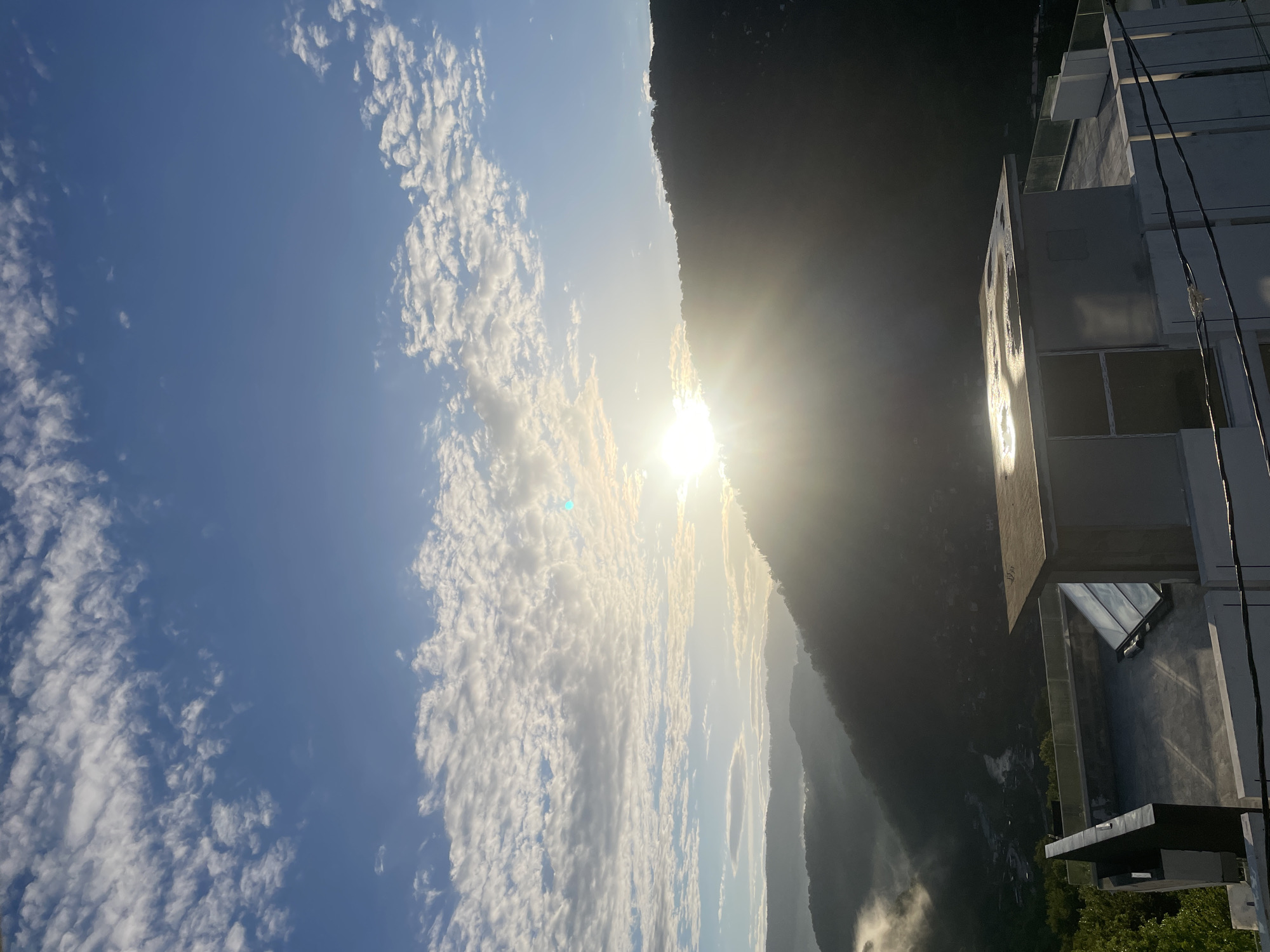 The image shows a daytime view with a bright sun partially obscured by clouds near a mountain range. The sky is mostly clear with scattered clouds. Part of a building is visible on the right side of the frame. It is impossible to verify the specific location in Baranovka, Russia from the image alone. There are no people or interactions visible.