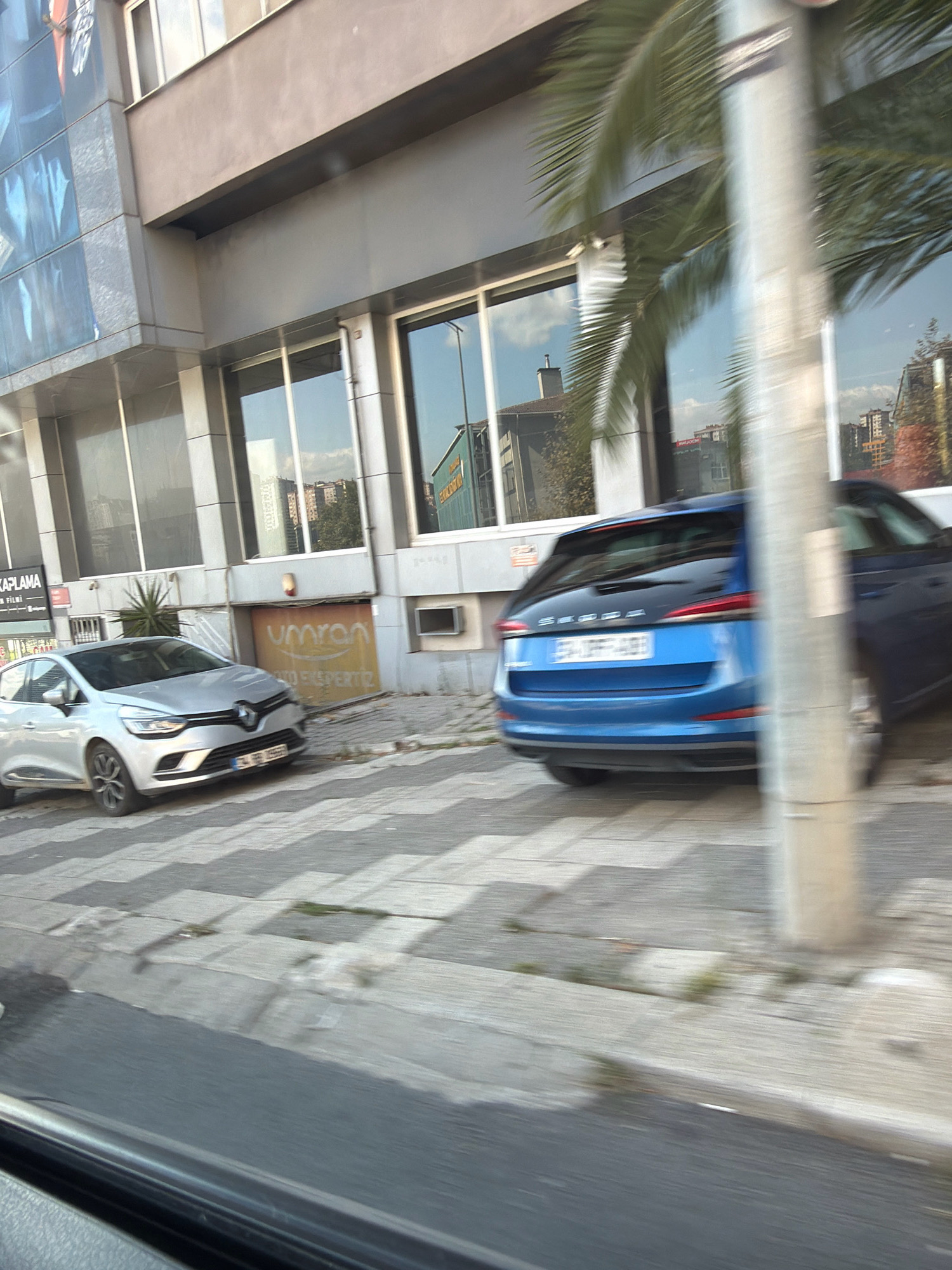 In Ataşehir, Turkey, a silver Renault Clio is parked at a crosswalk. To its right, a blue Skoda is parked. Both cars are positioned on a city street, adjacent to a building with a modern facade. A palm tree is visible, its fronds casting shadows on the building's windows. The building appears to be commercial, with large glass panes reflecting the sky and surrounding structures. A faded yellow sign with Turkish writing is affixed to the lower part of the building. The image captures a typical urban scene with vehicles and pedestrian infrastructure.