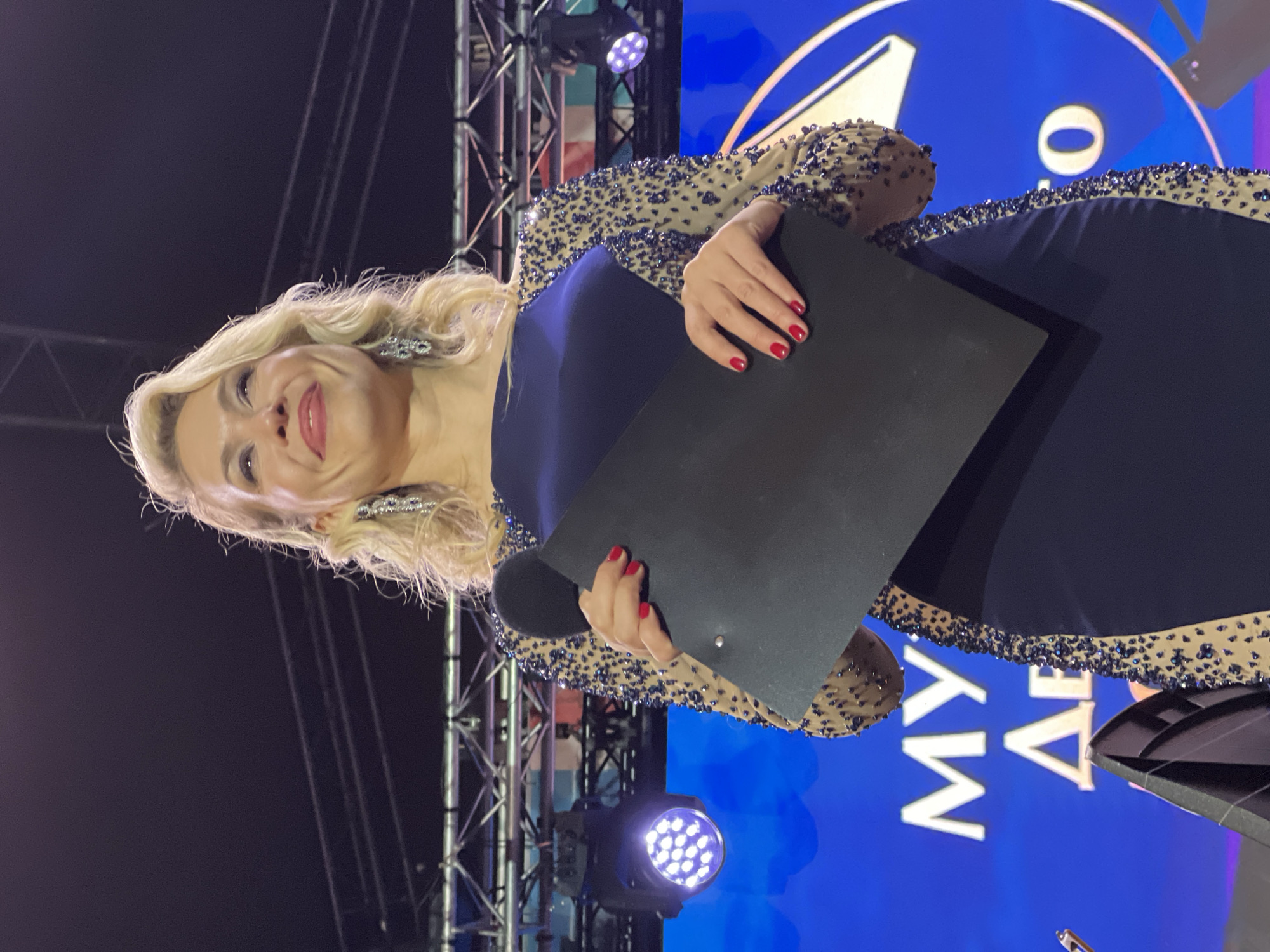 A woman with blonde hair, wearing a navy blue dress with beaded sleeves, is holding a black clipboard while standing on a stage. She has red nail polish on her fingernails. Behind her, there is stage lighting and a blue screen with white lettering. The stage is located in Saint Petersburg, Russia.