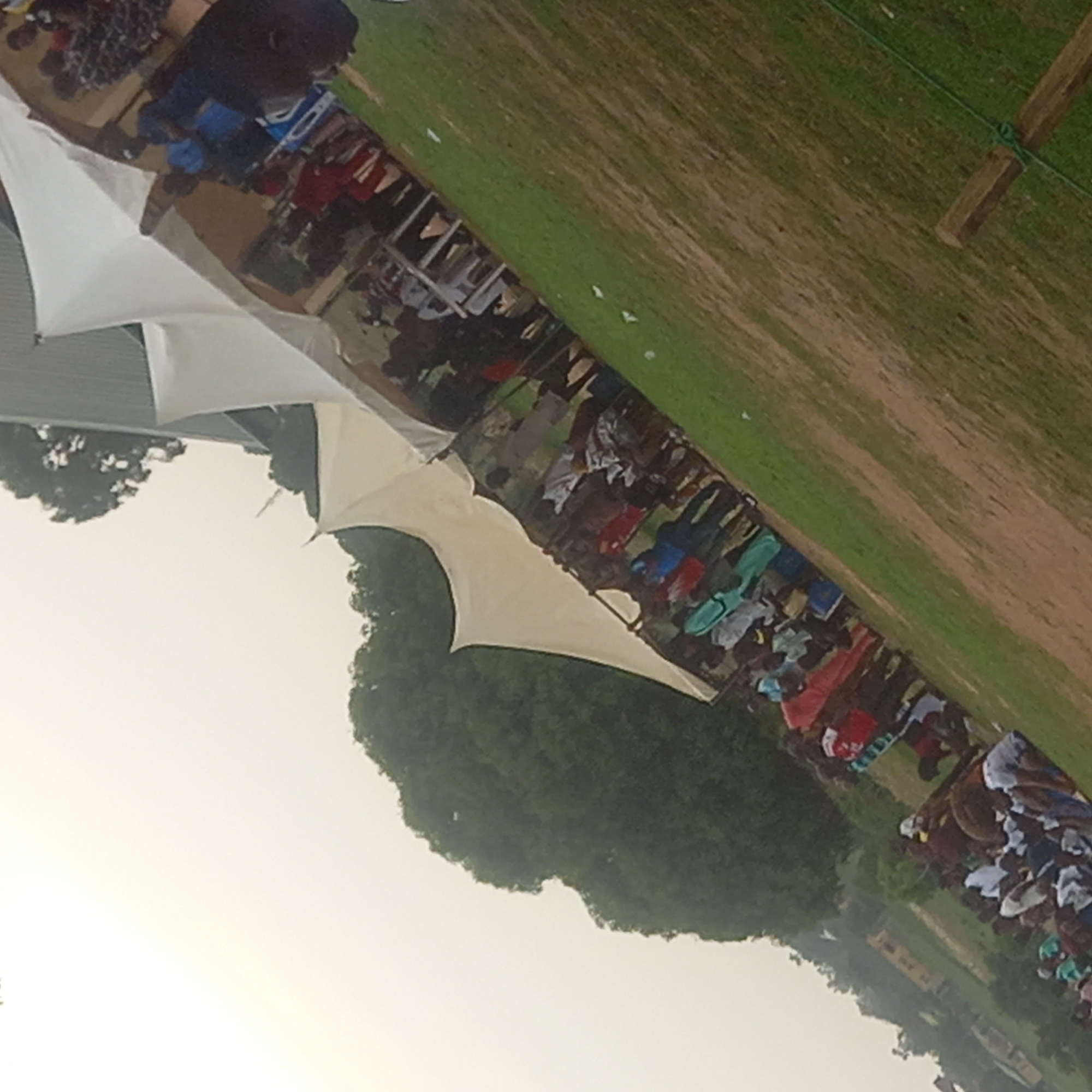 A ground-level, slightly angled shot captures a scene in Narayi, Nigeria. The environment consists of a grassy field with a light brown patch and a section of green grass. In the middle distance, a line of people is observed under white tents, seemingly engaged in some activity. The people are wearing various colors of clothing. Trees and a building are visible in the background. A wooden post is partially visible at the upper right. The sky appears light, suggesting daytime conditions.