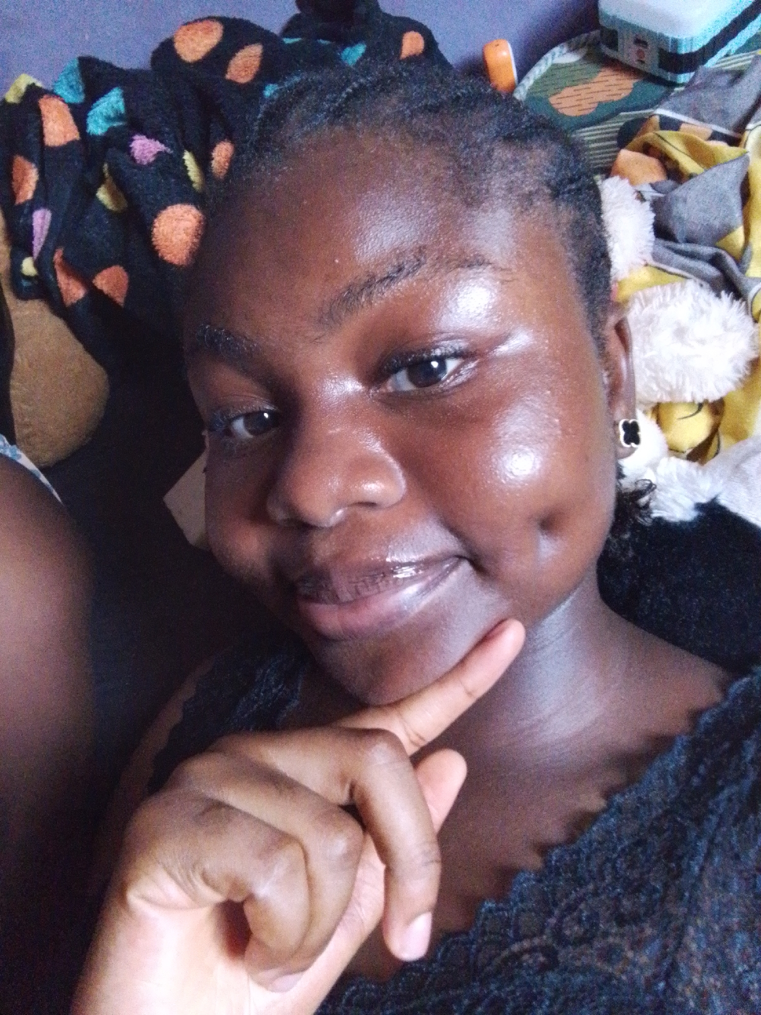 A dark-skinned young woman is lying down, her head surrounded by patterned bedding and a soft toy. She is smiling slightly with dimples visible on her cheeks and is wearing a black clover-shaped earring on her left ear. Her left hand is raised, with her index finger touching her cheek. The background is cluttered with various fabrics and objects, including what appears to be a plastic container. The location is unspecified, but presumed to be indoors in Nsukka, Nigeria.