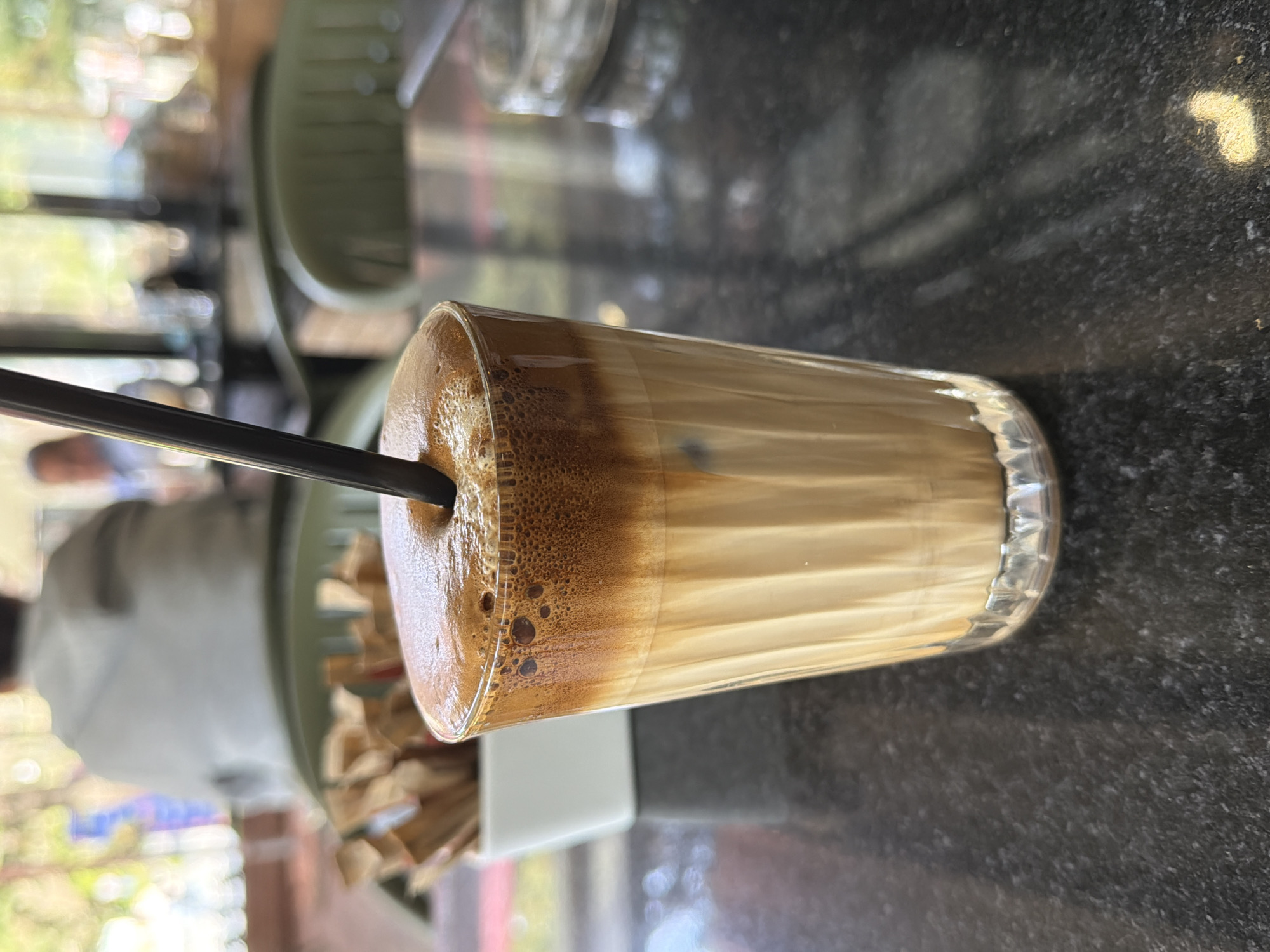 The image, rotated 90 degrees clockwise, presents a close-up view of a tall, clear glass of iced coffee, likely a frappe or Dalgona-style beverage, resting on a dark, speckled tabletop. A black plastic straw is inserted into the drink.
The coffee displays distinct layers: the top third of the glass is filled with a rich, dark brown, frothy foam, featuring visible bubbles. Below this, the beverage transitions into a creamy, lighter brown layer, further blending into a pale, milky or icy layer towards the bottom. The black straw extends downwards into these layers.
The setting appears to be a casual cafe in Stara Zagora, Bulgaria. The tabletop is a dark, possibly granite or marble, material with light speckles. To the left of the glass, partially visible in the blurred foreground, is a small white rectangular container, which might hold sugar packets or stirring sticks.
The background is significantly out of focus, indicating a shallow depth of field and drawing attention to the drink. Soft blurs of green suggest foliage or trees, implying an outdoor cafe or one with large windows overlooking greenery. Faint shapes of blurred furniture, possibly chairs, are also discernible, along with a vague reflection or figure of a person. The scene is brightly illuminated by natural light, consistent with daytime, suggesting weather suitable for a cold drink. There is no visible text in the image. The overall impression is one of relaxation and enjoying a refreshing beverage.