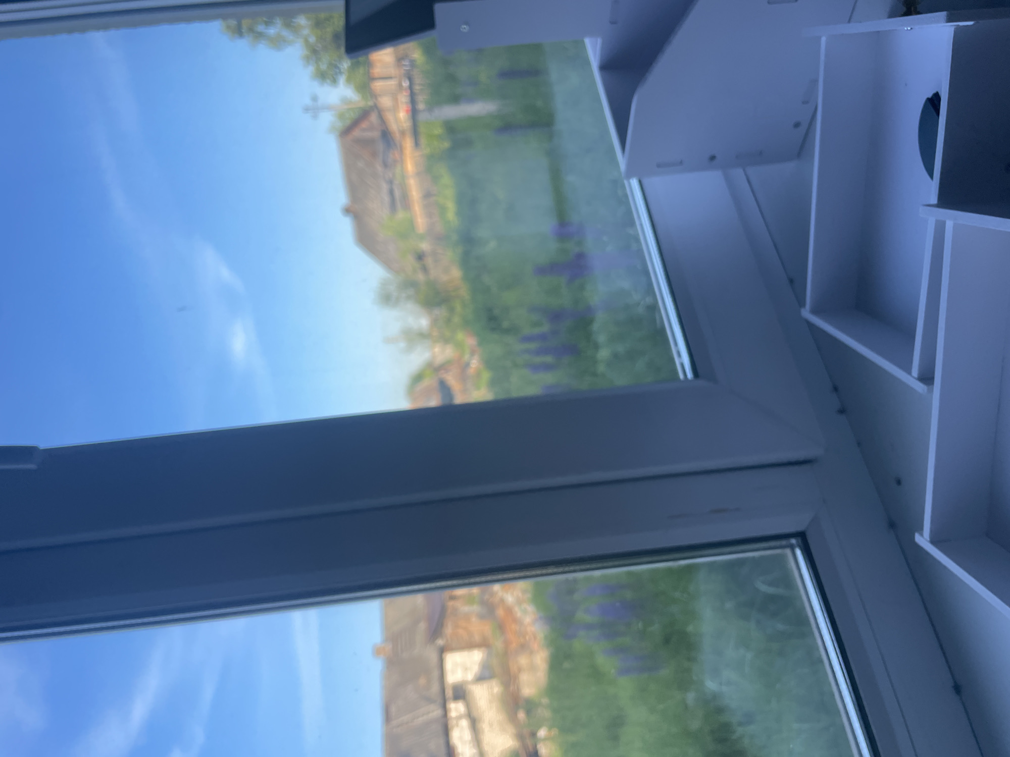 The image shows a view through a window of a rural landscape, likely in the vicinity of Krasnye Baki, Russia. The foreground is dominated by a white window frame and sill, with partially assembled white storage organizers on the sill. Outside the window, a green field with scattered purple flowers is visible, leading to a cluster of houses with traditional dark roofs. The sky is blue with thin, wispy clouds. The overall impression is of a bright, sunny day in a rural setting.