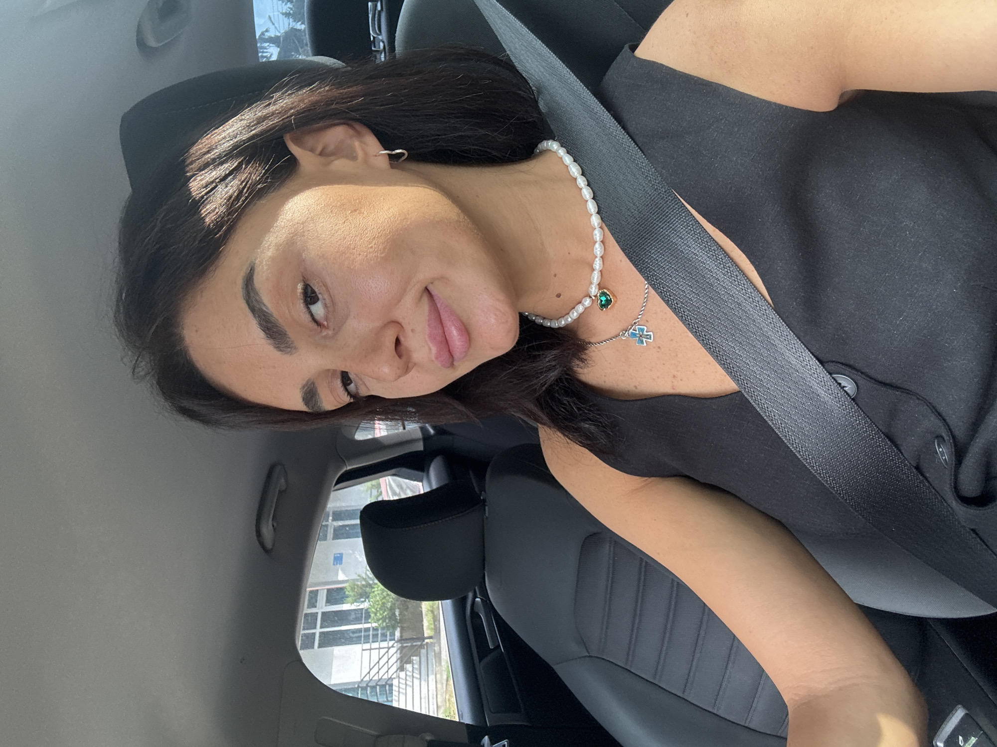 A woman with dark hair and light skin is sitting in the driver's seat of a car, facing the camera. She is wearing a black V-neck top and a pearl necklace with a green heart-shaped pendant and a silver cross pendant. She is wearing a seatbelt. The interior of the car is visible, including the black leather seats and a portion of the dashboard. Outside the car, a building with windows and trees can be seen. The scene appears to be in Tbilisi, Georgia, based on the architecture.