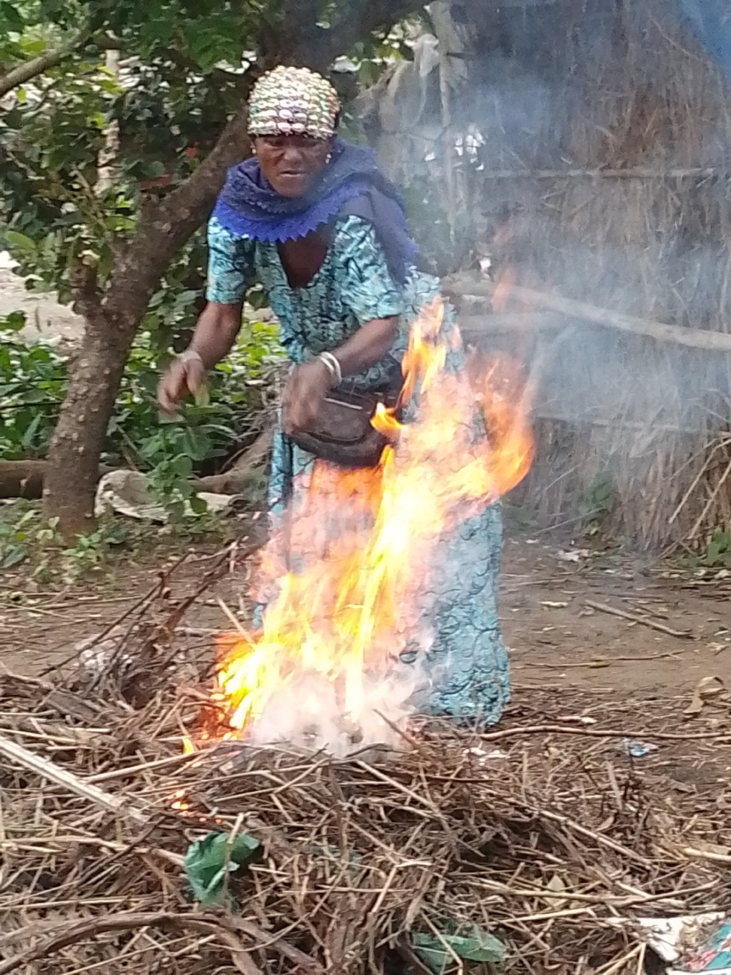 An elderly African woman wearing a blue patterned dress and a blue headscarf with a beaded cap is standing in front of a pile of burning dry vegetation. She is holding a dark bag with her right hand, and her left arm is extended towards the fire. A blue scarf is draped around her neck. The scene takes place outdoors in a rural setting, possibly in Otulu, Nigeria, with trees and foliage in the background. The ground is covered with dry leaves and twigs. Smoke rises from the fire, obscuring some of the background.