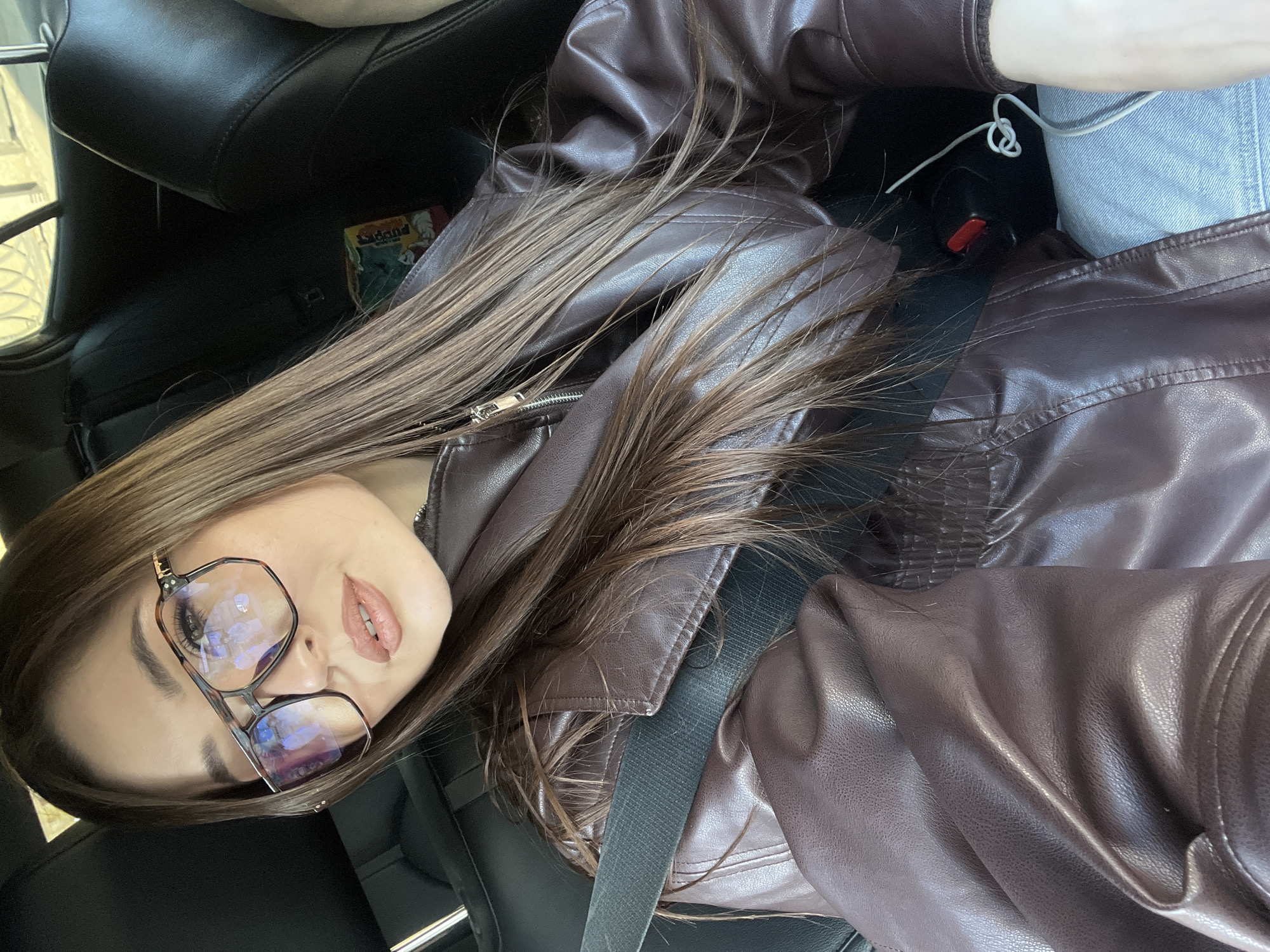 A young woman is photographed in the interior of a car. She wears glasses and a brown leather jacket, and a black seatbelt crosses her chest. Part of her hair falls across her jacket. The car seats are black leather. A charging cable is visible in the upper right of the frame, plugged into a red and black device. A piece of packaging, green, yellow, and white in color, is partially visible between the woman and the car seat. Based on the appearance of the woman and the high-quality vehicle interior, the location could be a contemporary car in an urban area, but there is no specific information to confirm the location as Moscow, Russia.
