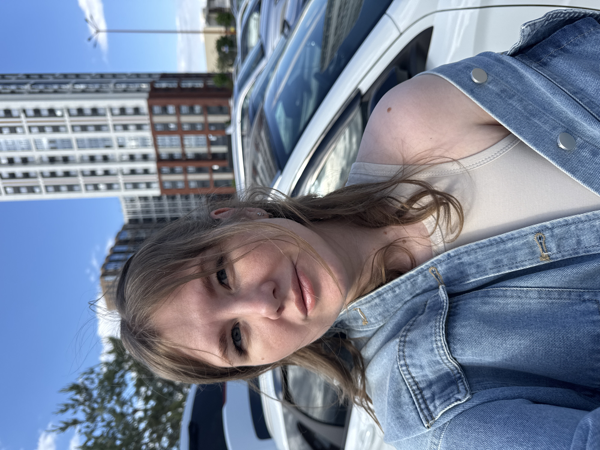 A young woman is taking a selfie outdoors. She is wearing a beige tank top and a light blue denim jacket. In the background, there are parked white cars and a tall building with a mix of white and brown facade. The sky is blue. Based on the building's architecture and information available, the location is likely in the city of Perm, Russia.