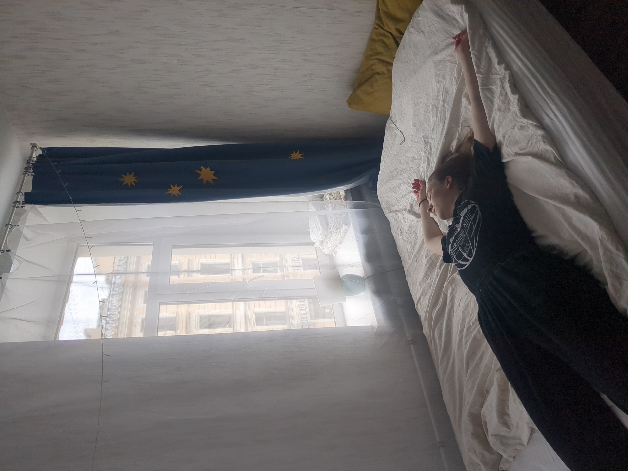 A medium-shot, high-angle photograph depicts a bedroom in Saint Petersburg, Russia. A young woman lies on a bed covered with a wrinkled, beige sheet. She wears a black t-shirt and pants. She stretches her right arm upward. A yellow pillow is on the bed near her head. The window is covered with a sheer, white curtain and white mosquito netting. A string of lights hangs near the window. The window faces a building with ornate facades and windows. A blue banner with gold star decals hangs above the window. The ceiling is white with subtle texture.