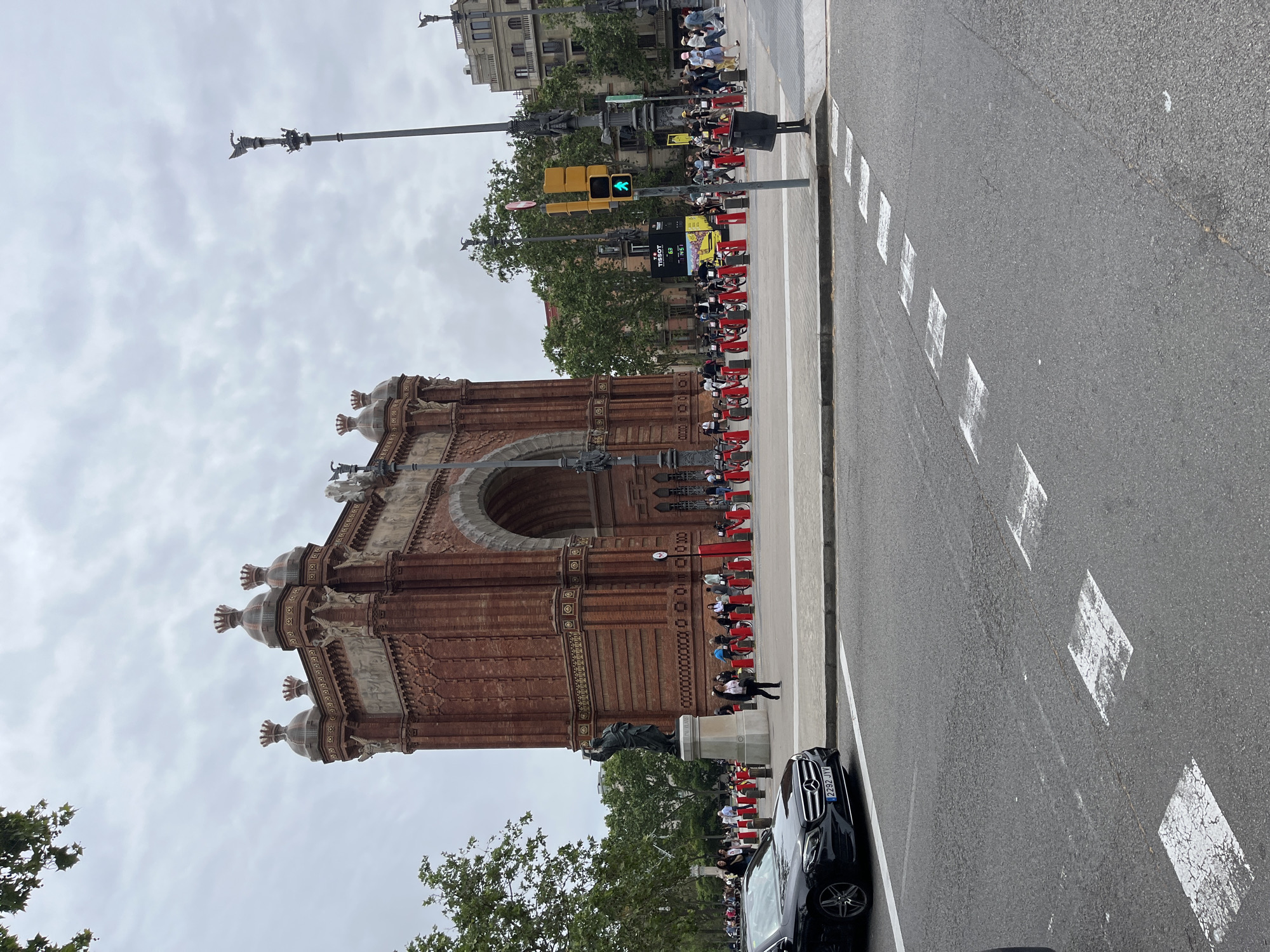 The image captures a scene near the Arc de Triomf in Barcelona, Spain, under a cloudy sky. The Arc de Triomf, a brick monument, stands prominently on the left. A long line of people, many seated on red benches or barriers, stretches along the walkway in front of the arch. Some people are walking. A black Mercedes car is parked on the edge of the road. The road is grey with white diamond markings. A traffic light shows green for pedestrians. Street lamps with sculptures on top and buildings are visible in the background.