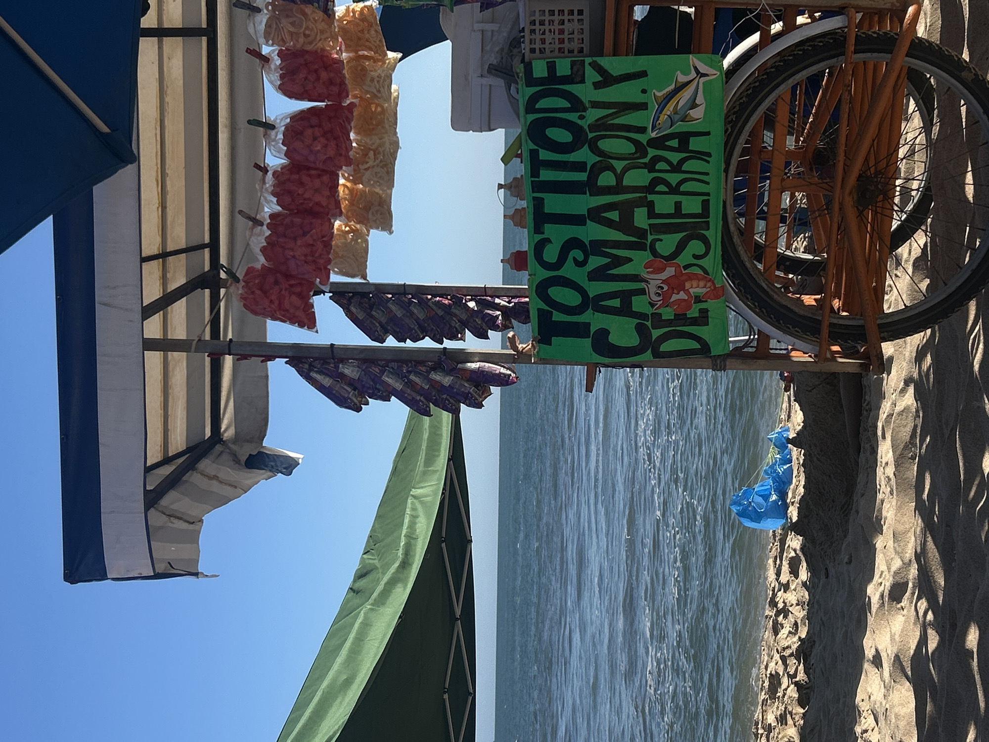 The scene depicts a food cart on a beach in Mazatlán, Mexico. The cart, likely mobile, is adorned with various bags of chips or snacks hanging from its frame, alongside a sign advertising "Tostitos de Camaron y de Sierra" (Shrimp and Sierra Tostitos) with cartoon images of a fish and a shrimp. A bicycle wheel is attached to the cart, suggesting its mobility. A green awning and a blue and white striped canopy provide shade over the cart. The environment shows a sandy beach meeting the ocean, under a clear blue sky. A blue plastic bag is discarded on the beach near the cart.