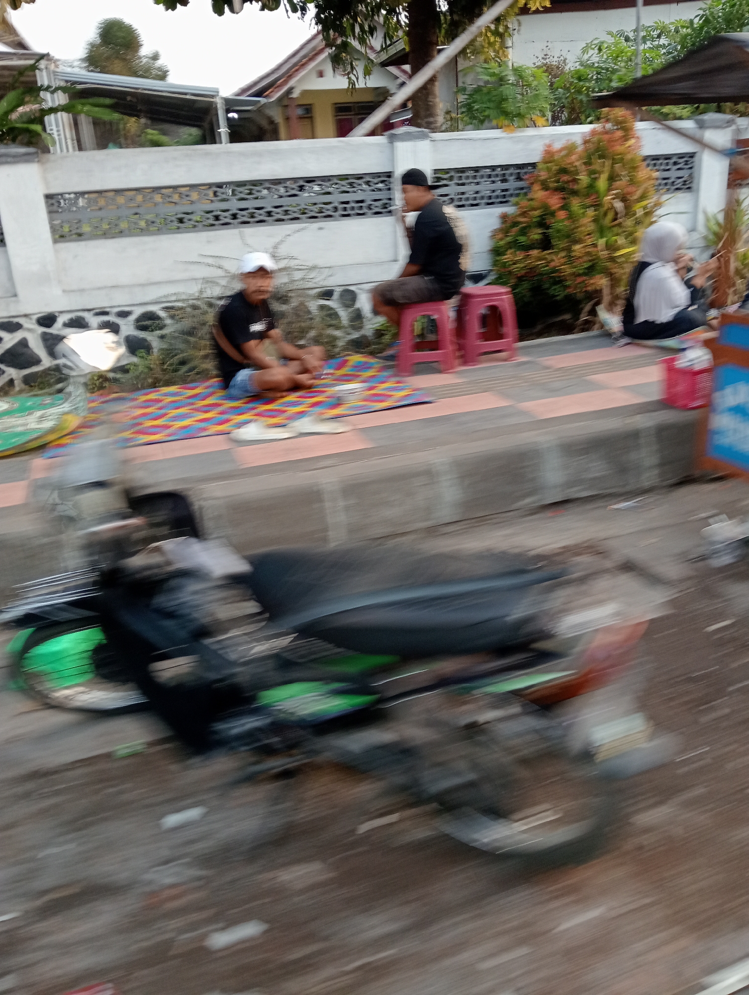 The image depicts a street scene in Grabag, Indonesia, with a blurred motion effect suggesting movement. In the background, a house with a light-colored facade and a tiled roof is visible behind a low white wall topped with decorative metalwork. Adjacent to the house, a colorful, bushy plant with orange and green foliage is situated near the wall.
In the middle ground, three individuals are present. One person, wearing a white cap and black t-shirt, sits on a patterned mat on the ground. To their right, another person, dressed in a black t-shirt and shorts, is seated on a red stool. Further to the right, a person wearing a white hijab and dark clothing is also seated on the ground. The ground area features a tiled pathway.
In the foreground, a motorcycle is prominently featured, also exhibiting motion blur. The motorcycle has a black seat and appears to have green accents on its wheels and body. The overall scene is likely an outdoor public space, possibly a roadside or sidewalk area in Grabag.