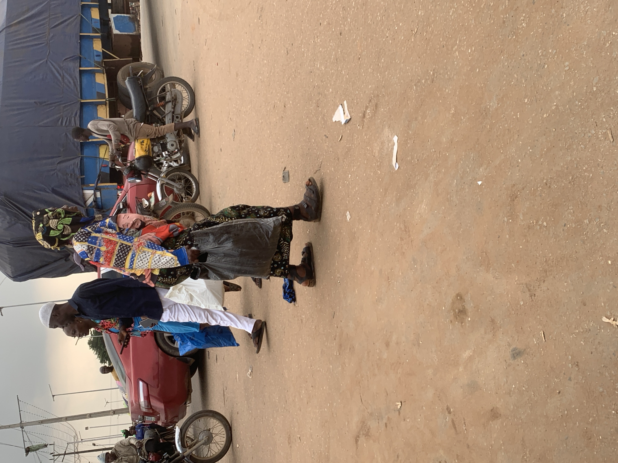 A street scene in Shagamu, Nigeria, features a variety of people and vehicles. In the foreground, a woman wearing a colorful patterned dress and a headscarf walks to the right, carrying a black plastic bag. To her left, a man in a blue shirt and white pants walks towards the left, holding a smartphone. Behind them, a red sedan is parked. Further back, a man sits on a motorcycle, and a large blue truck with a tarp-covered load is visible. The ground is dusty and appears to be a road surface. The sky is overcast.