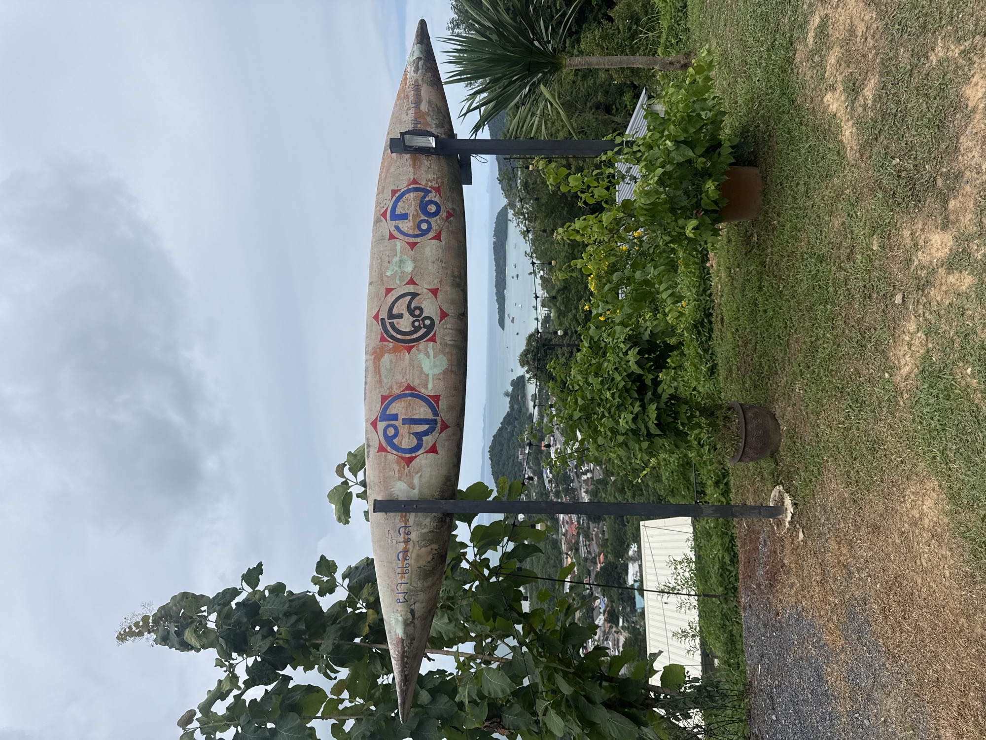A long, curved object, resembling a boat or a large seed pod, is mounted horizontally on two dark metal posts in the foreground of the image. The object is painted with light colors and features several circular designs. On the left side, there are blue and red circular symbols with radiating lines. Towards the center and right, there are black circular symbols with intricate patterns. Faded text is visible on the object's surface. The scene appears to be an outdoor viewpoint overlooking a coastal town. In the background, there is a body of water with several boats and islands. The sky is overcast with gray clouds. Lush green trees and vegetation are present in the foreground and on the hillsides. Two potted plants, one on the left and one on the right, flank the central object. A pathway of gravel and dry grass is visible in the lower left of the image. This location is a viewpoint in Wichit, Thailand, offering a panoramic view of the coastline and surrounding landscape.