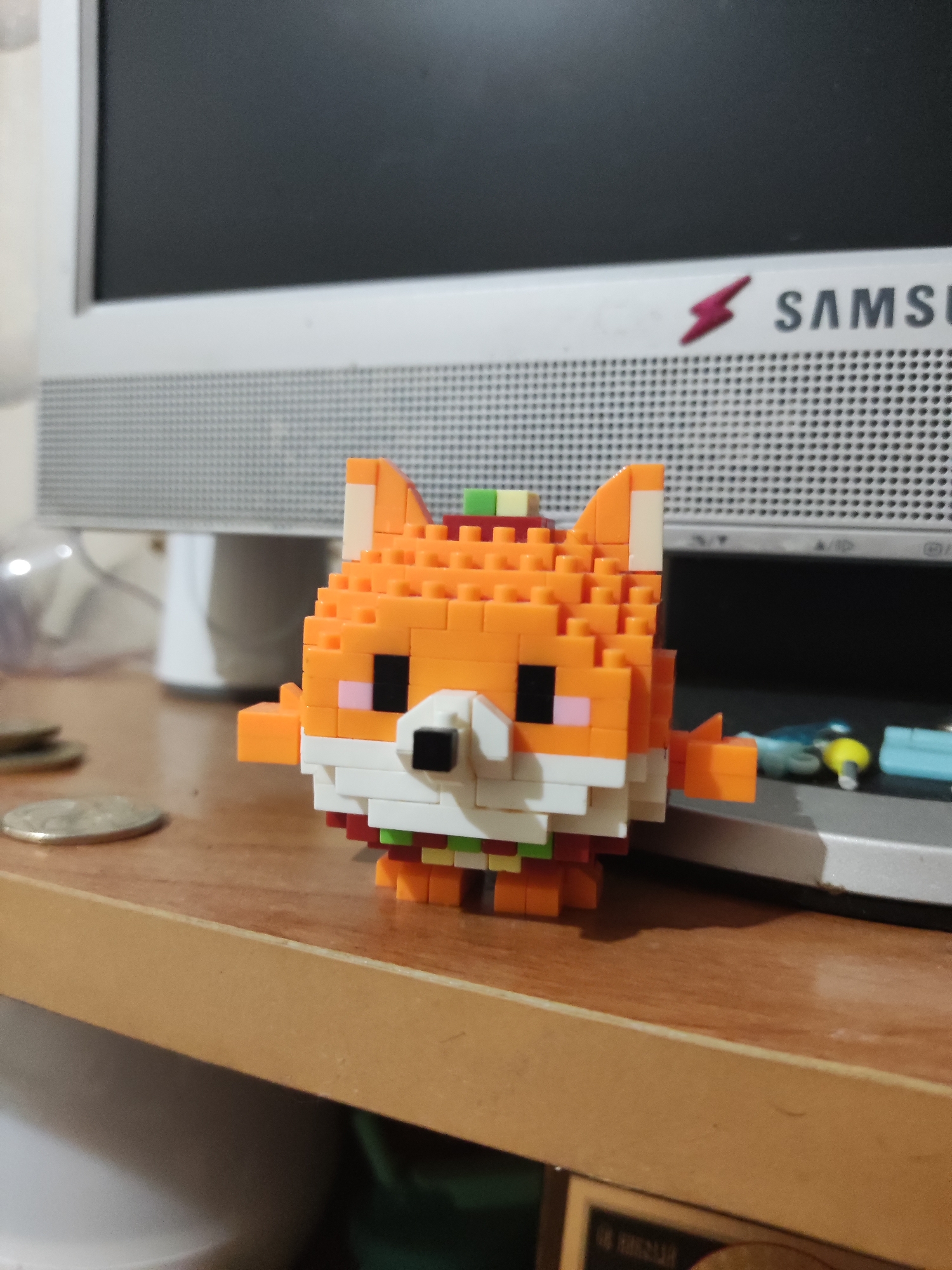 A pixelated, orange fox figurine made of building blocks sits on a wooden desk in front of a computer monitor. The figurine has white and black block details for its face and colorful blocks for its accessories. Two coins are visible on the desk to the left of the figurine. The computer monitor behind the figurine has a silver frame and a dark screen. The overall environment suggests an indoor setting, likely a workspace or a home office. There is no indication of the city of Shymkent, Kazakhstan.