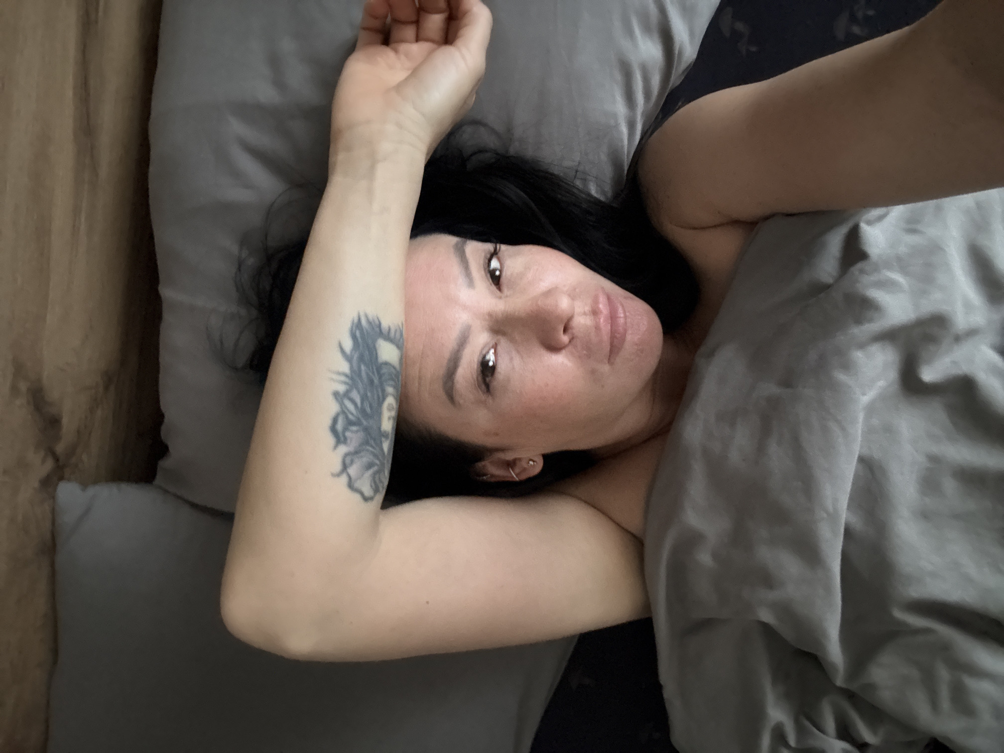 A woman with dark hair lies in a bed with gray linens. She has a tattoo on her left arm. The background includes a wooden surface. The location information is not discernible from the image.