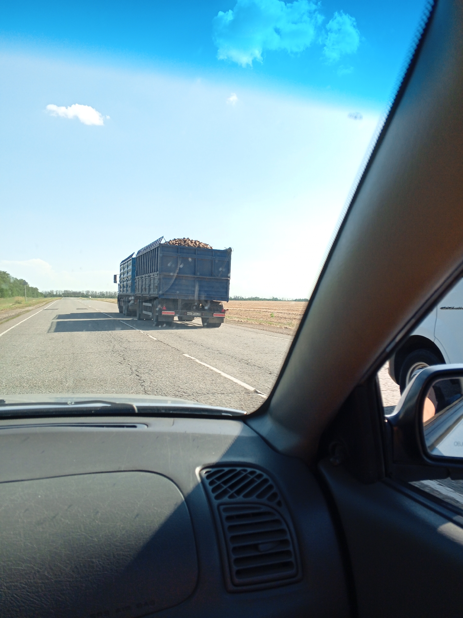 A blue truck, loaded with produce, drives down a two-lane road under a clear blue sky with white clouds. The truck's trailer is large and appears to be made of metal. The road is asphalt and has white lane markings. To the right of the road, there is a field of dry crops. The image is taken from inside a vehicle, looking out through the windshield. The dashboard and part of the interior of the vehicle are visible in the foreground. The scene appears to be a rural area.
This image was taken on the M4 highway near Vyselki, Russia. The M4 highway is a major federal highway in Russia that runs from Moscow to Novorossiysk. Vyselki is a rural settlement in Krasnodar Krai, Russia. The area is characterized by agricultural land and is a common route for agricultural transport.