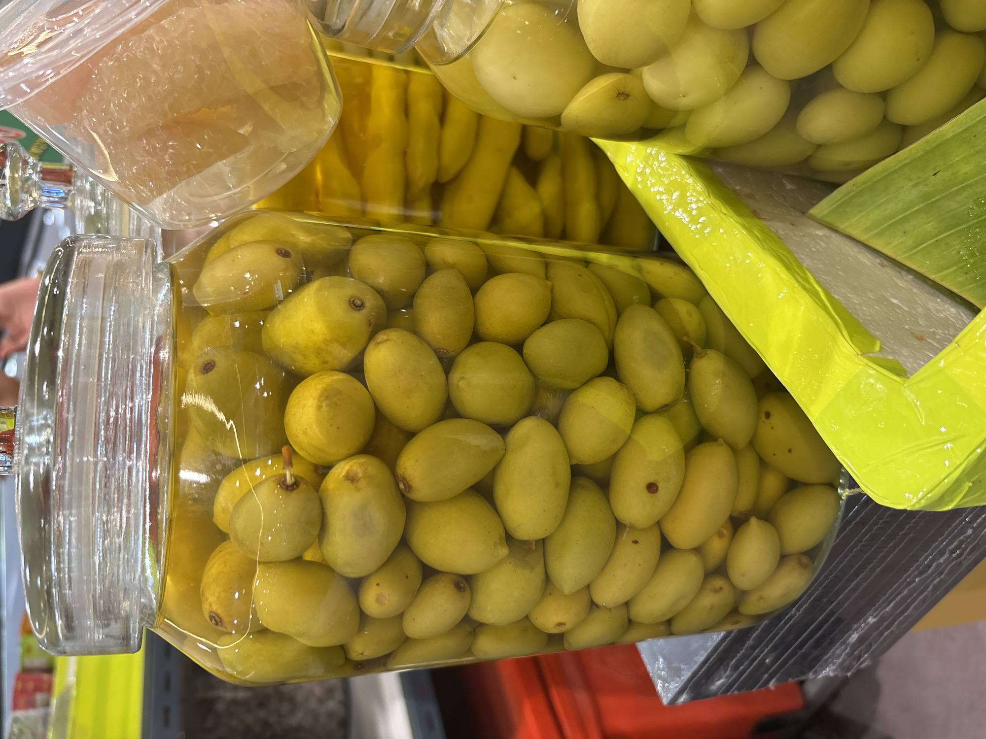 The image shows three glass jars filled with yellow-green fruit preserved in liquid. The jars are stacked on top of each other on a surface, likely a table or display. The scene appears to be indoors, possibly in a shop or market, located in บ้านบางรักษ์, Thailand.
